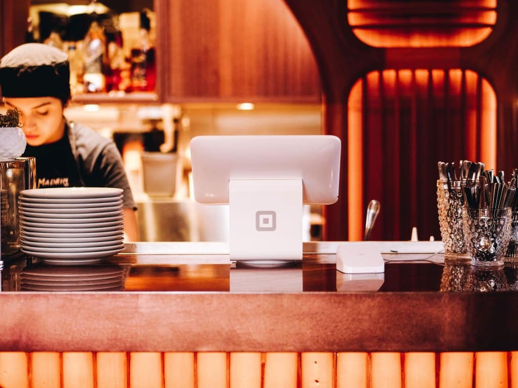 La barra de un restaurante con un terminal de punto de venta de Square; a la izquierda, platos apilados; a la derecha, cubiertos en sus soportes; y, al fondo, una persona trabajando bajo una iluminación cálida.