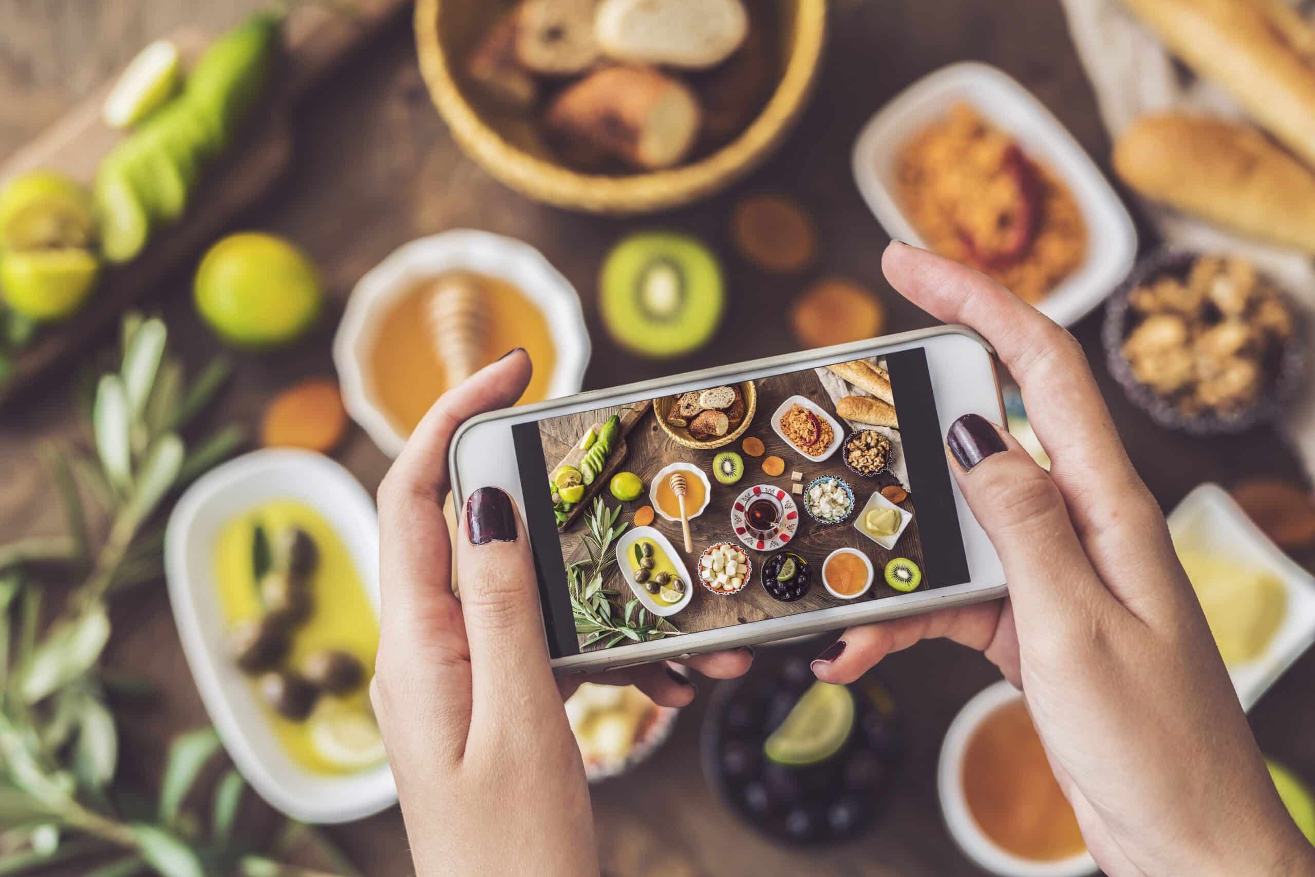 Una persona sostiene un smartphone para hacer una foto de una mesa repleta de diversos aperitivos, salsas para mojar, fruta, pan y salsas, vista desde arriba.
