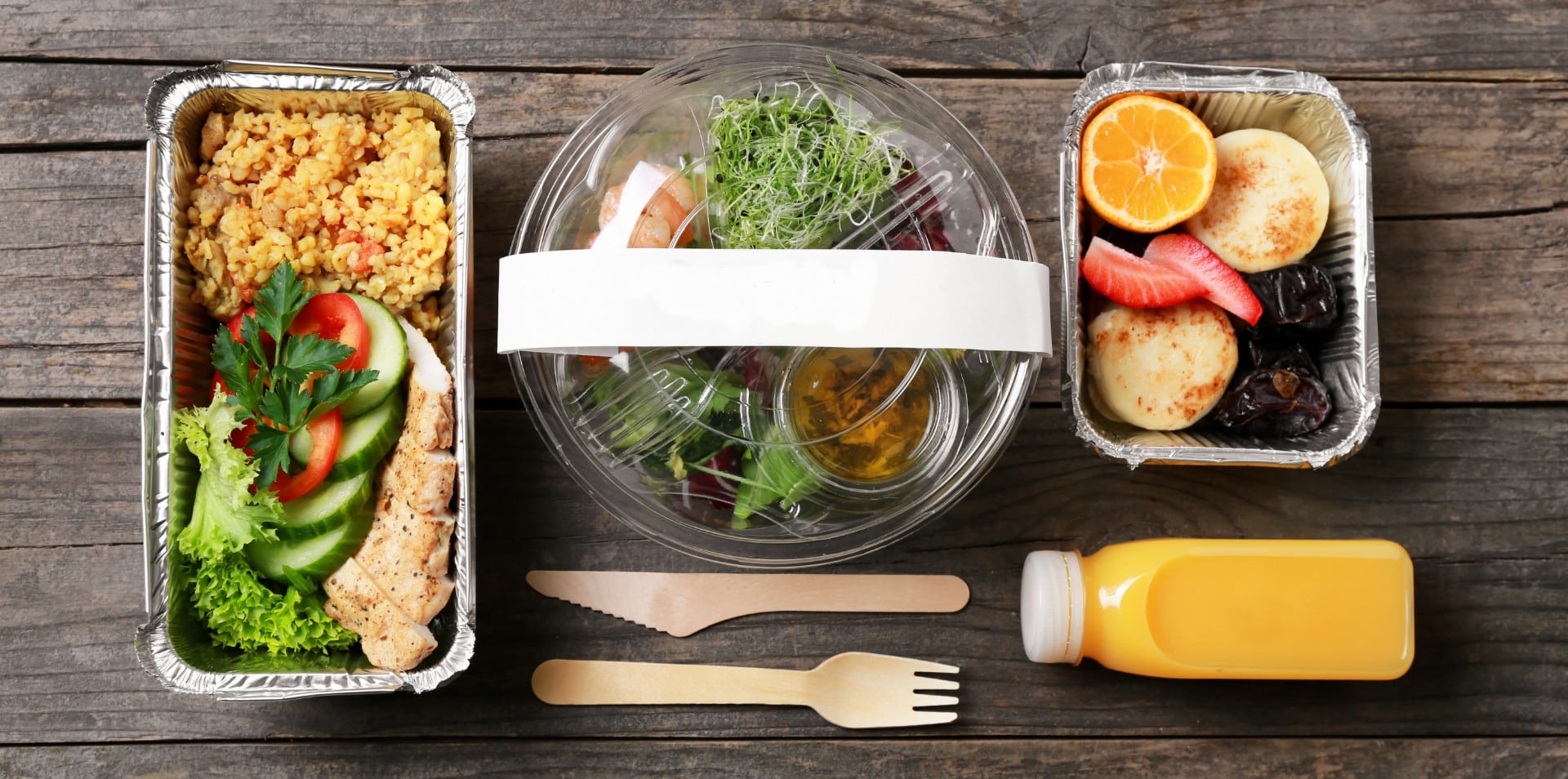 Una comida saludable con ensalada, pollo a la parrilla, arroz, frutas variadas, tortitas, una botella de zumo y cubiertos de madera, todo ello dispuesto sobre una mesa rústica de madera.