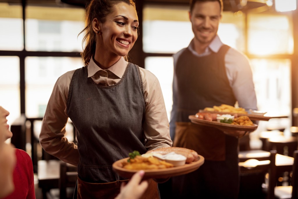 Dos camareros, ataviados con delantales, sirven platos de comida —entre los que se incluyen patatas fritas, hamburguesas y ensalada— a los clientes de un restaurante con ventanas iluminadas por el sol al fondo.