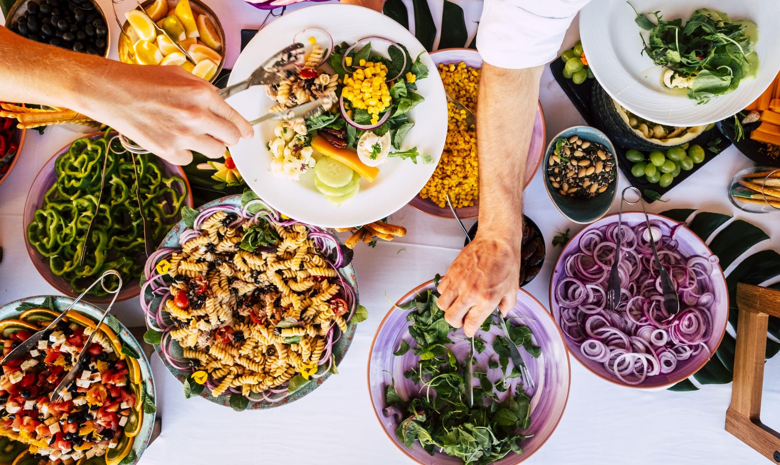 Vista desde arriba de una colorida barra de ensaladas con cuencos de ensalada de pasta, verduras de hoja verde, cebolla cortada, maíz, hortalizas y fruta. Dos personas se sirven ellas mismas, cogiendo los alimentos con unas pinzas y con las manos.