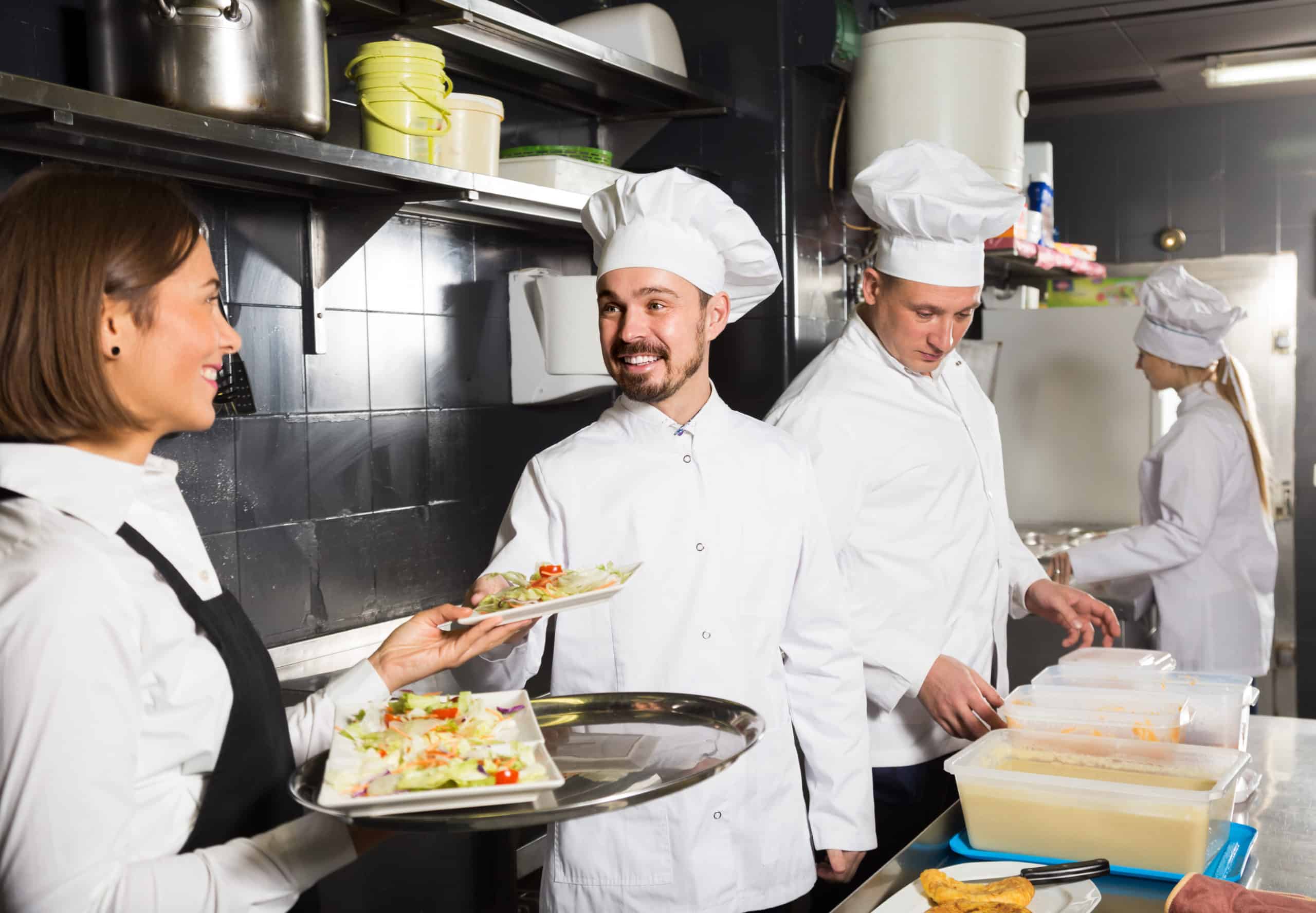 Cuatro cocineros en una cocina industrial: dos de ellos sonríen y charlan mientras sostienen platos con comida, y los otros dos preparan platos en la barra, al fondo.
