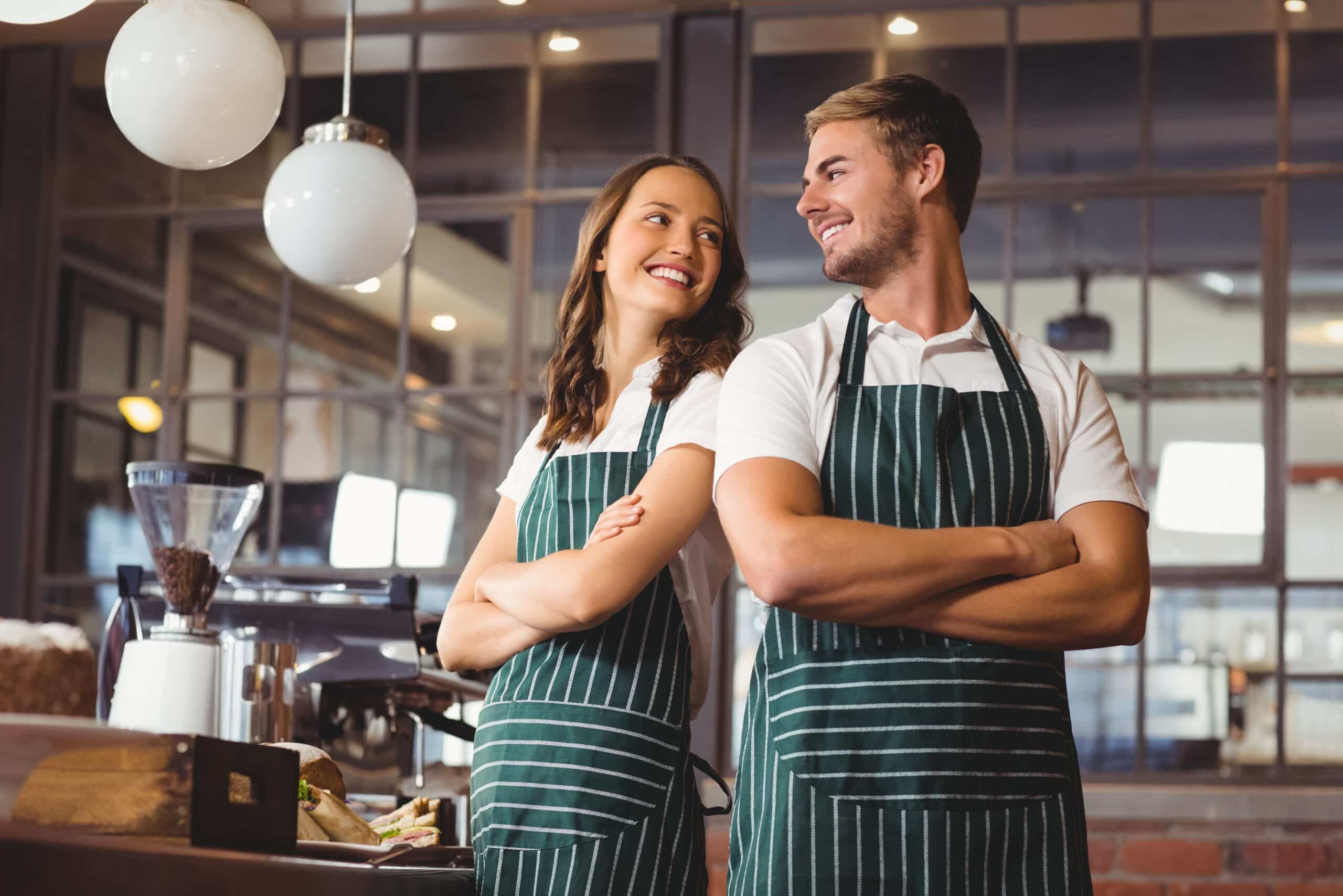Dos empleados de una cafetería, que llevan delantales a rayas verdes, están de espaldas el uno al otro, sonriéndose mutuamente en una cafetería con iluminación moderna y grandes ventanales al fondo.
