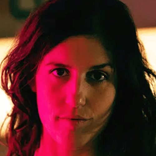 Se muestra un primer plano de una persona con el pelo largo y oscuro, iluminada de forma dramática con luz roja en un lado del rostro y luz natural en el otro, con un fondo interior desenfocado.