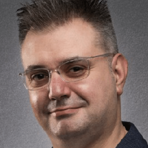 Un hombre de pelo corto y oscuro, con gafas y barba bien recortada, vestido con una camisa oscura, sonríe levemente sobre un fondo gris.