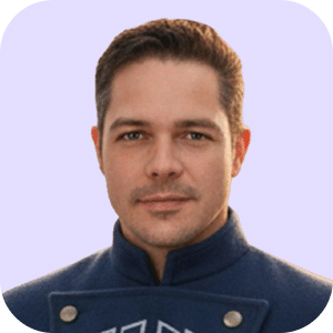 Un hombre con el pelo castaño corto, una ligera barba incipiente y un abrigo azul de cuello alto está de pie delante de un fondo de color morado claro.
