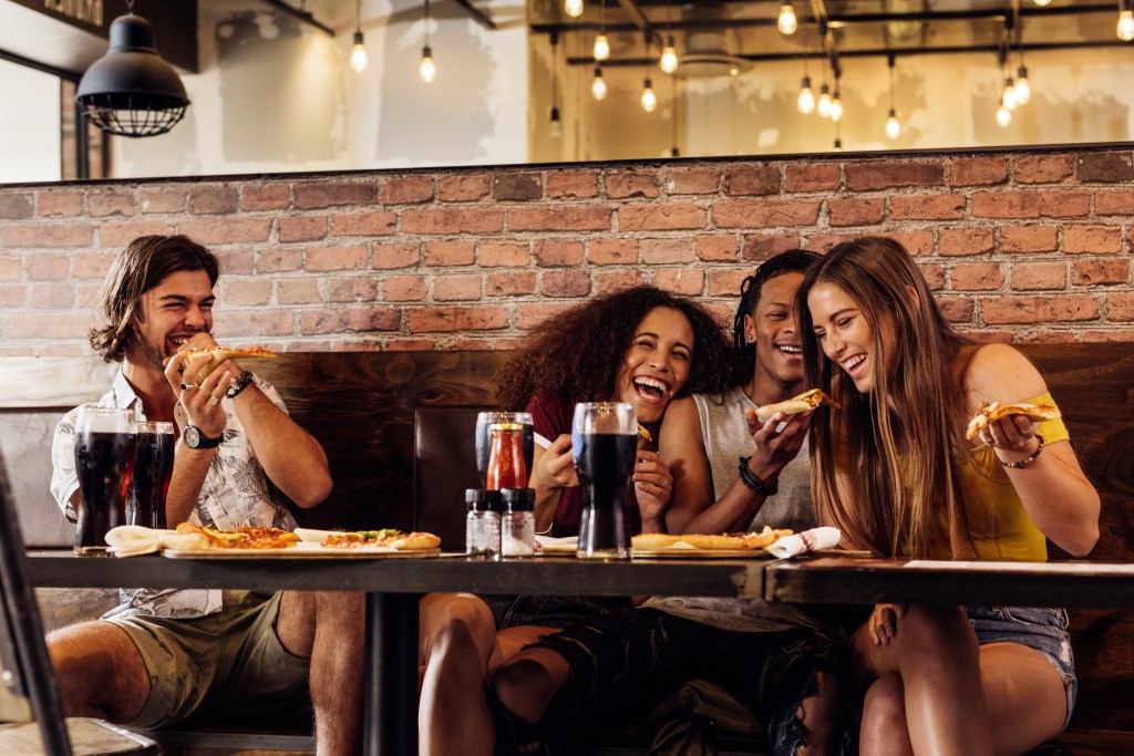 Cuatro amigos están sentados en una mesa de un restaurante, riendo y comiendo pizza juntos. Sobre la mesa hay refrescos y condimentos, con una iluminación cálida y una pared de ladrillo al fondo.