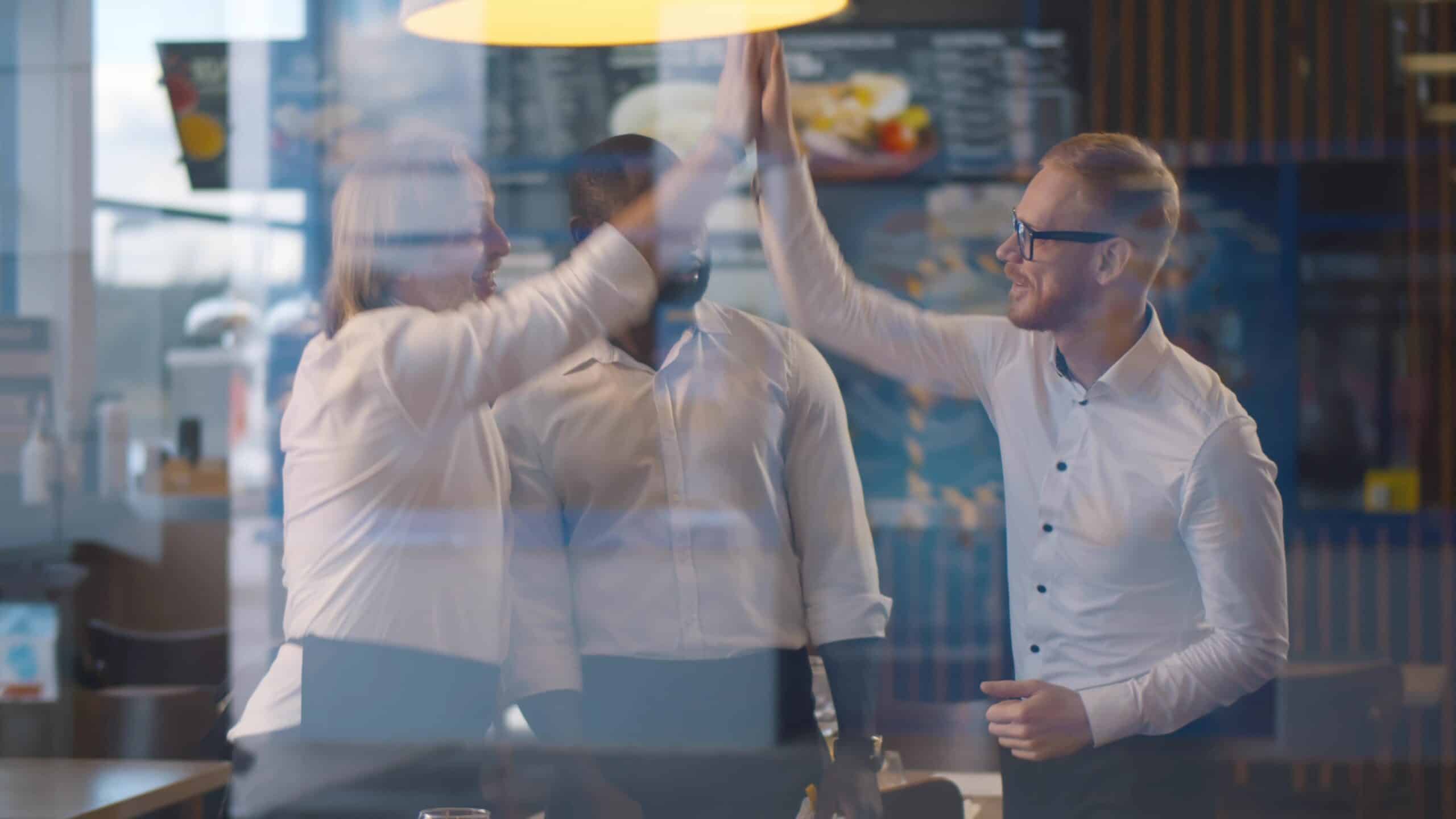 Tres personas con camisetas blancas se dan un choque de manos en un restaurante, de pie y sonriendo, con un cartel con el menú visible al fondo y reflejos en la ventana de enfrente.