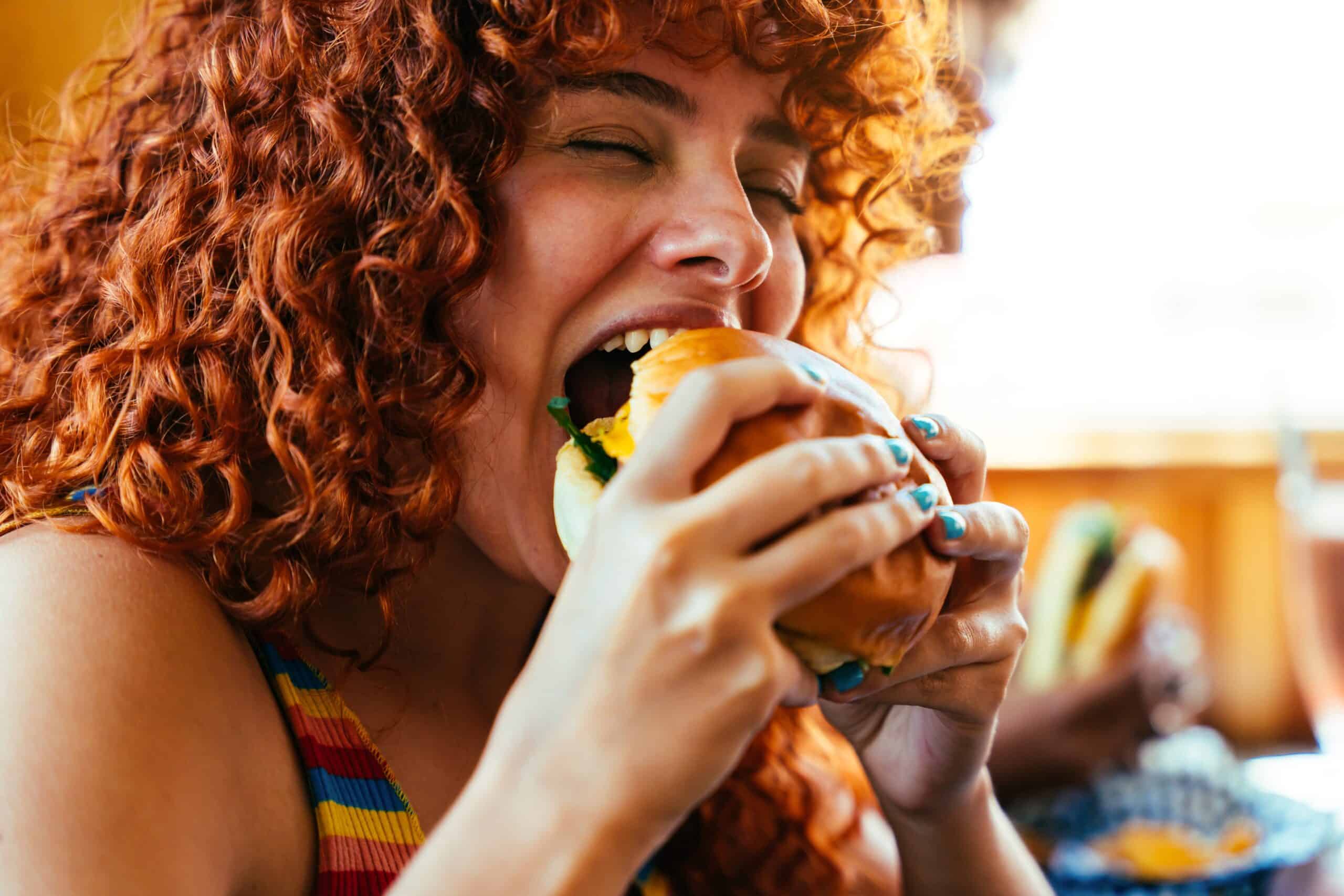 Una mujer de pelo rojo y rizado sonríe mientras le da un buen mordisco a un bocadillo; lleva una camiseta de rayas de colores y disfruta de su comida en un ambiente luminoso e informal.