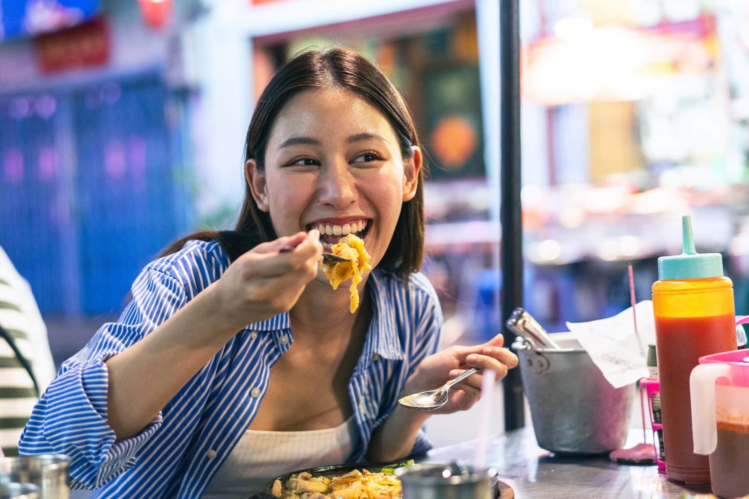 Una mujer con una camiseta a rayas azules sonríe mientras come fideos con un tenedor en un puesto de comida callejero; se ven condimentos de colores y, al fondo, un mercado difuminado.