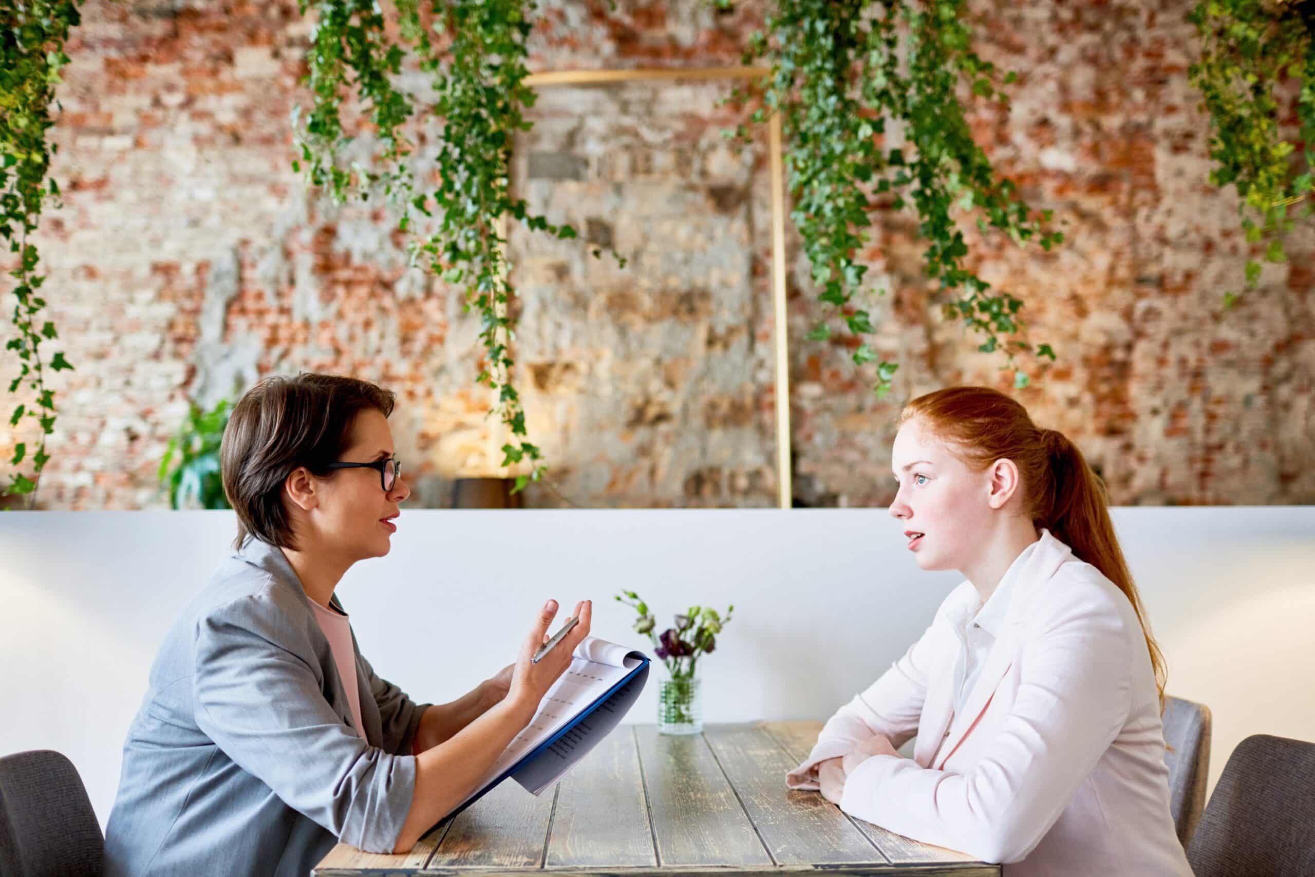 Dos mujeres están sentadas una frente a la otra en una mesa de madera, en un espacio moderno decorado con plantas, y mantienen una conversación. Una de ellas sostiene un cuaderno, y ambas parecen concentradas y atentas.