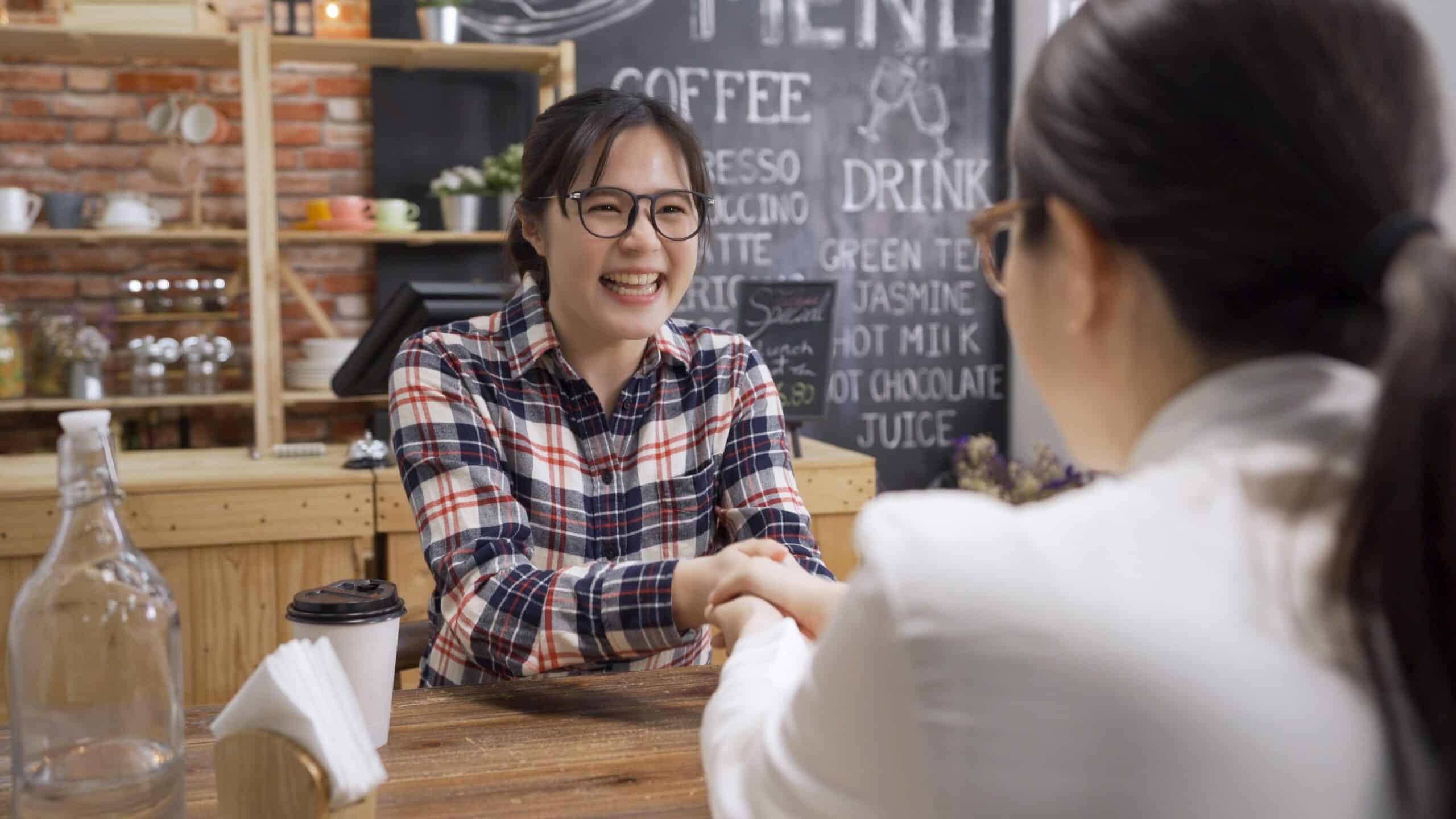 Dos mujeres están sentadas una frente a la otra en la mesa de una cafetería, sonriendo y dándose la mano. Al fondo se ve una pizarra con el menú, junto con una taza de café y una botella de cristal sobre la mesa de madera.