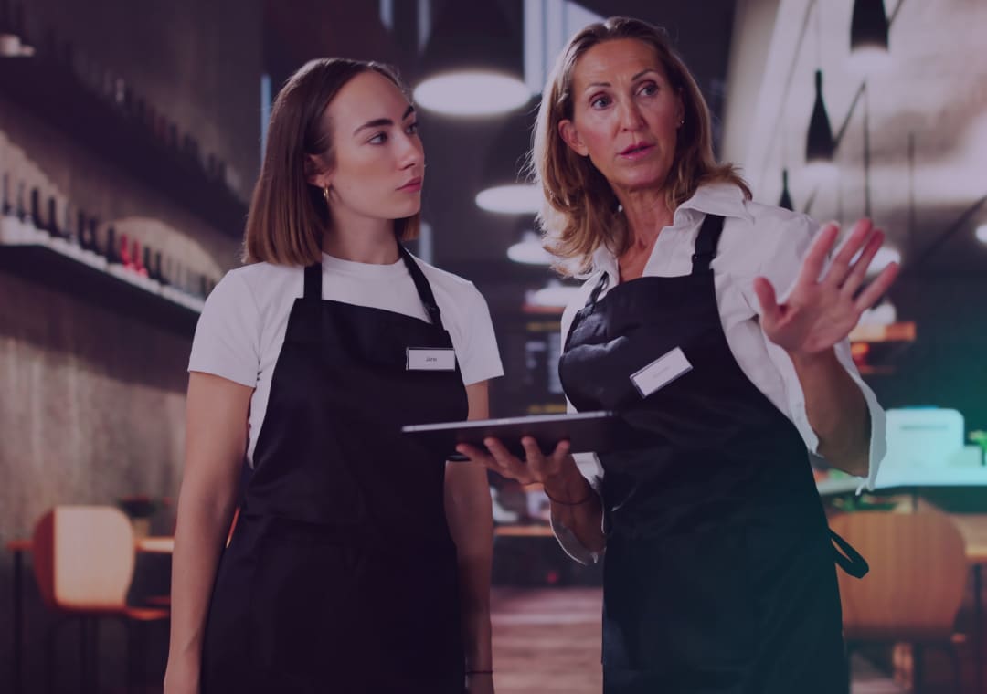Dos mujeres con delantales negros están de pie en un restaurante moderno. Una de ellas sostiene una tableta y gesticula, como si le estuviera explicando algo a la otra, que la escucha con atención. Al fondo se ven estanterías con botellas y mesas vacías.