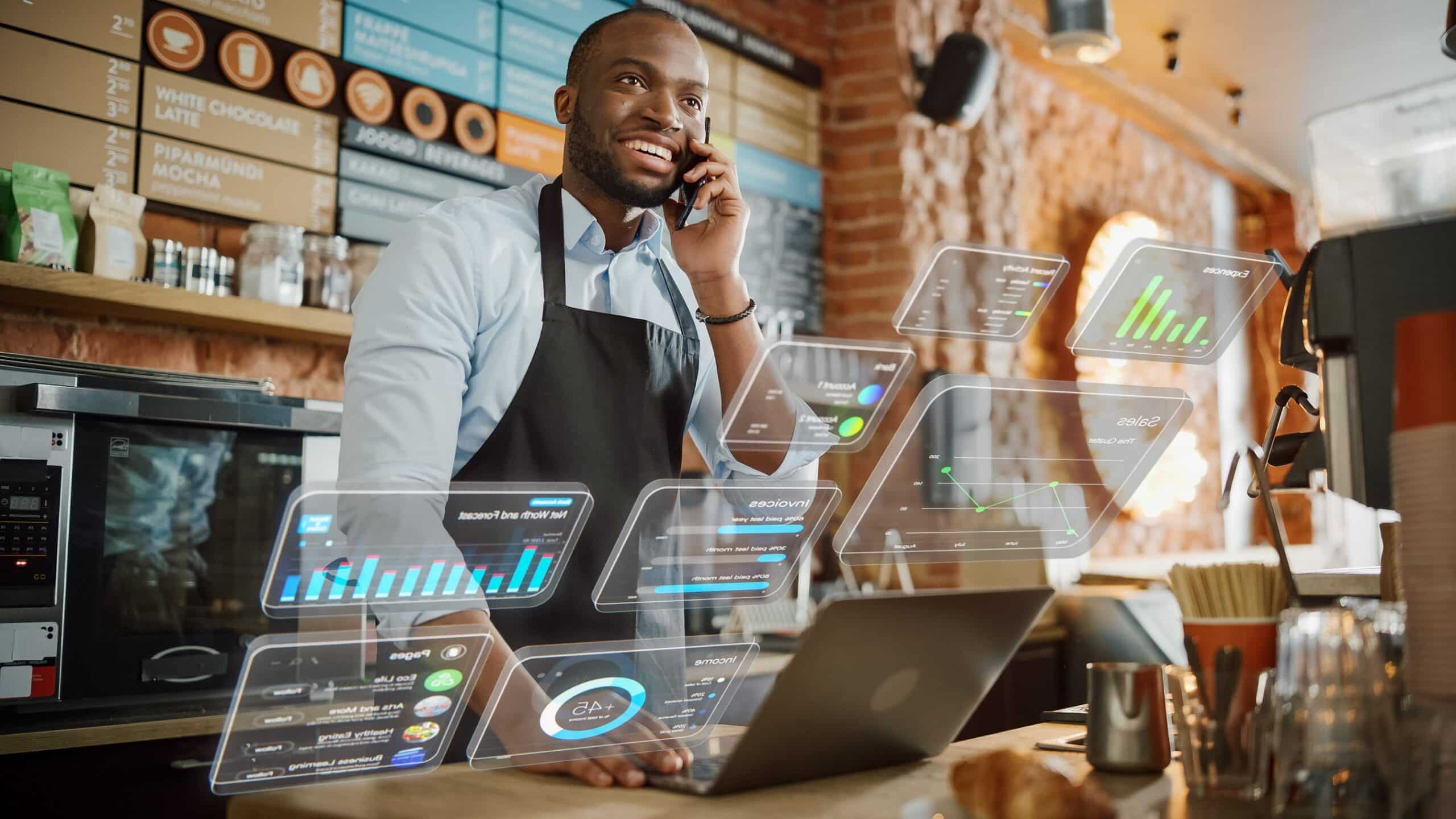 Un barista sonriente, con delantal, habla por teléfono detrás del mostrador de una cafetería, con un ordenador portátil abierto y gráficos y tablas de datos digitales superpuestos delante de él, lo que sugiere que se dedica al análisis empresarial o a la integración tecnológica.