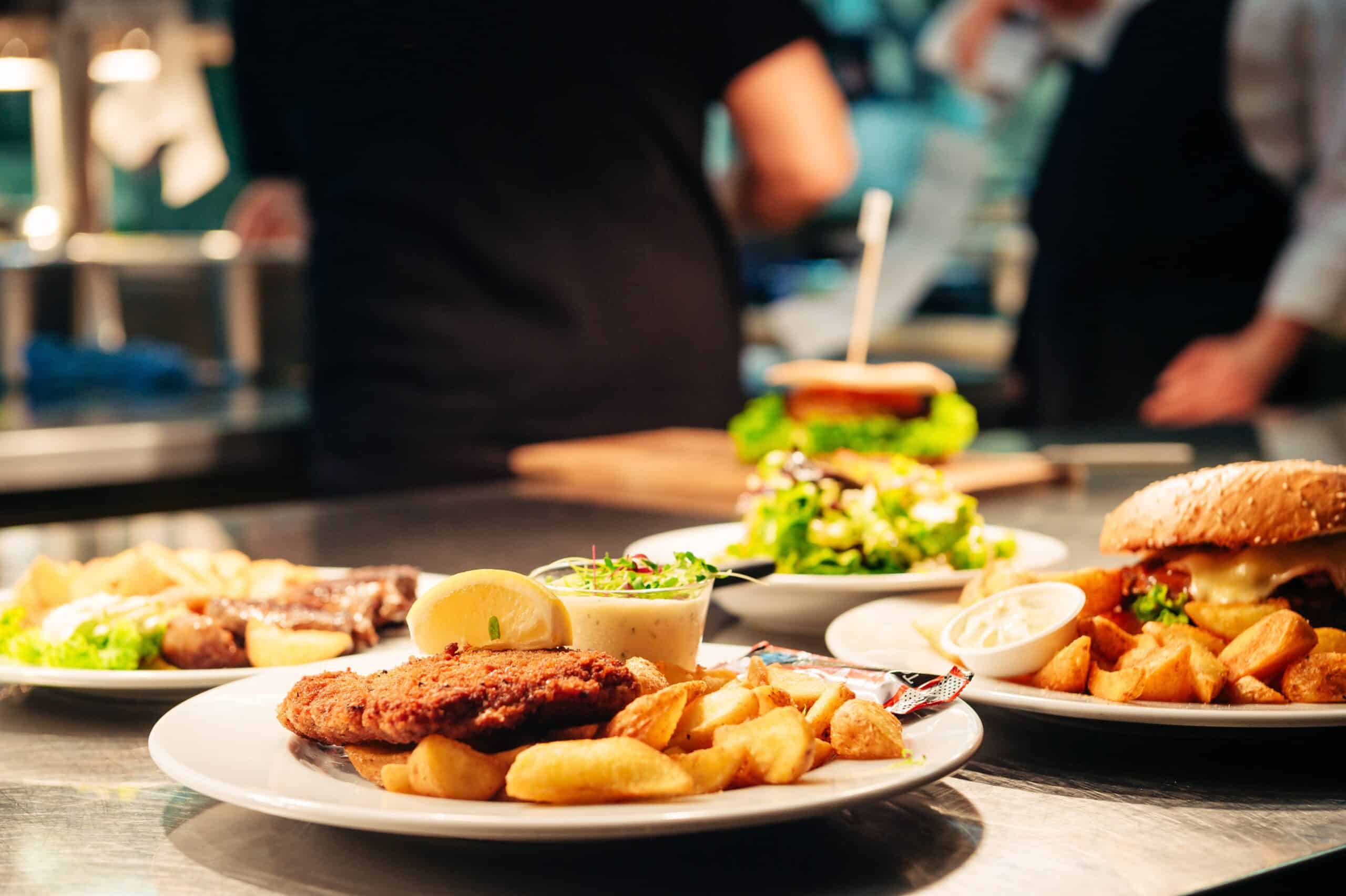 Un primer plano de platos con comida frita, gajos de patata, ensalada y hamburguesas en la barra de un restaurante, con personas desenfocadas y el fondo de la cocina.