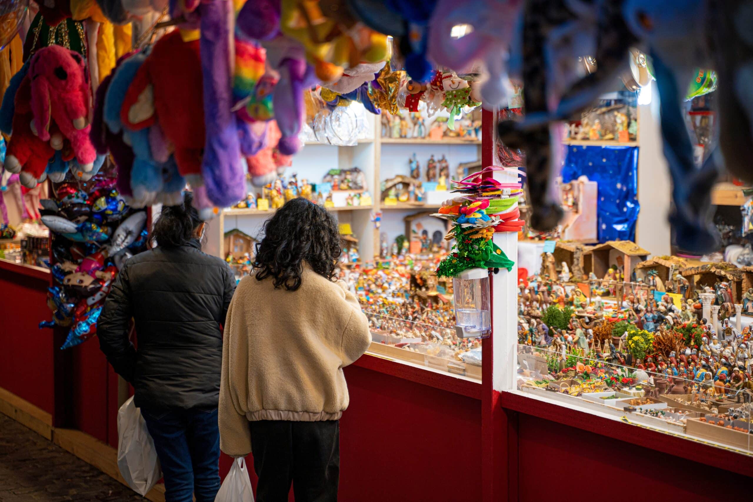 Dos personas están de pie junto a un colorido puesto de mercado repleto de figuritas y peluches, echando un vistazo a los artículos expuestos. La escena es animada y festiva, con peluches colgados y una decoración llamativa.