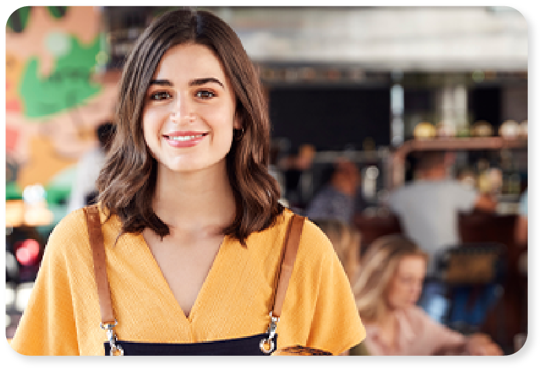 Una mujer con el pelo castaño a la altura de los hombros, vestida con una camiseta amarilla, sonríe a la cámara en el animado ambiente de una cafetería, con personas borrosas al fondo.