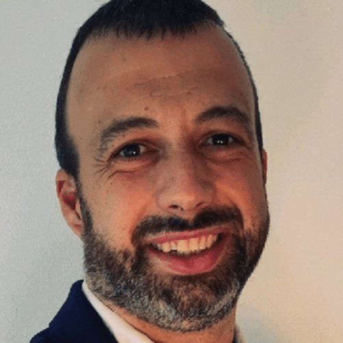 Un hombre de pelo corto y oscuro y con barba sonríe a la cámara. Lleva una chaqueta oscura y una camisa blanca, y está de pie frente a un fondo liso de color claro.