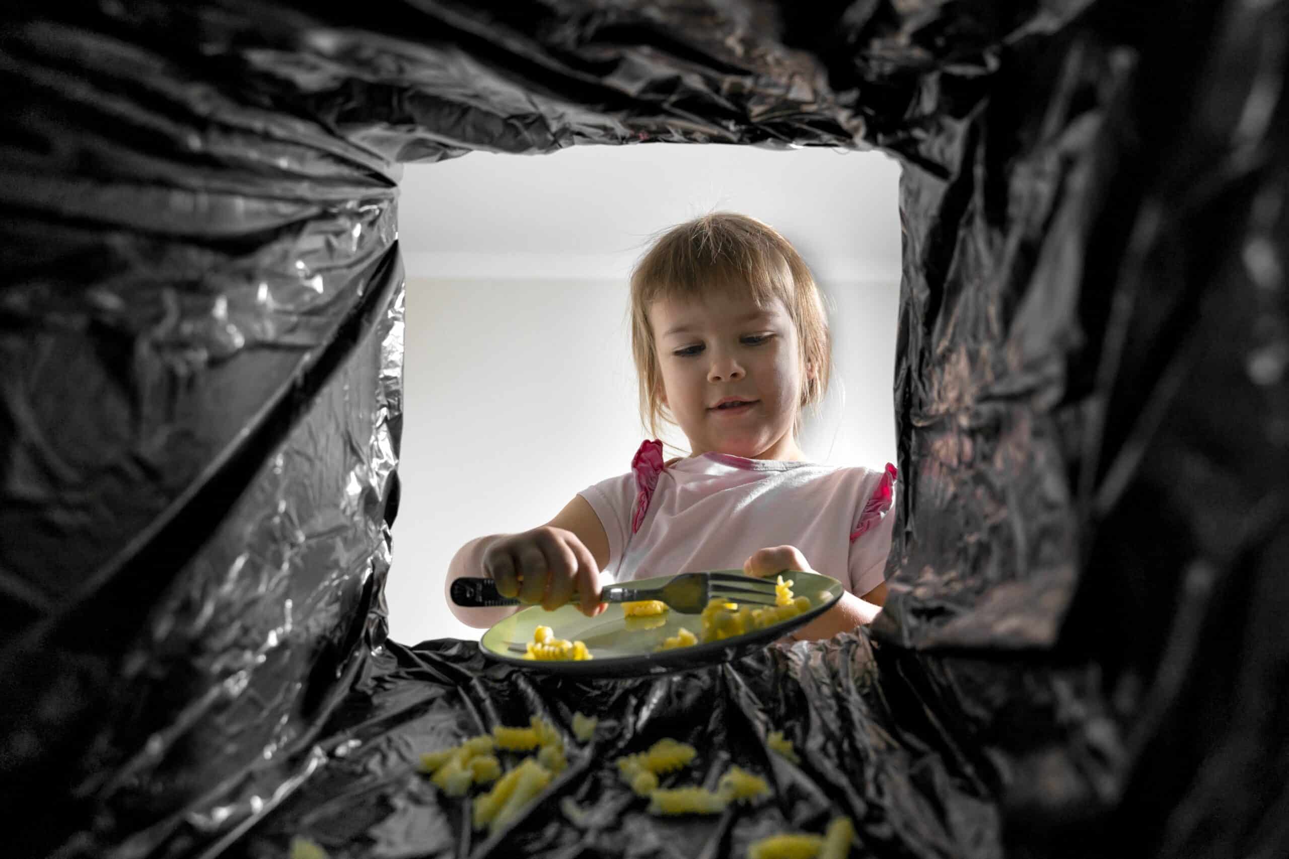 Un niño recoge los restos de comida de un plato y los echa en una bolsa de basura negra; la imagen se ve desde el interior de la bolsa, en la que caen trocitos de pasta.
