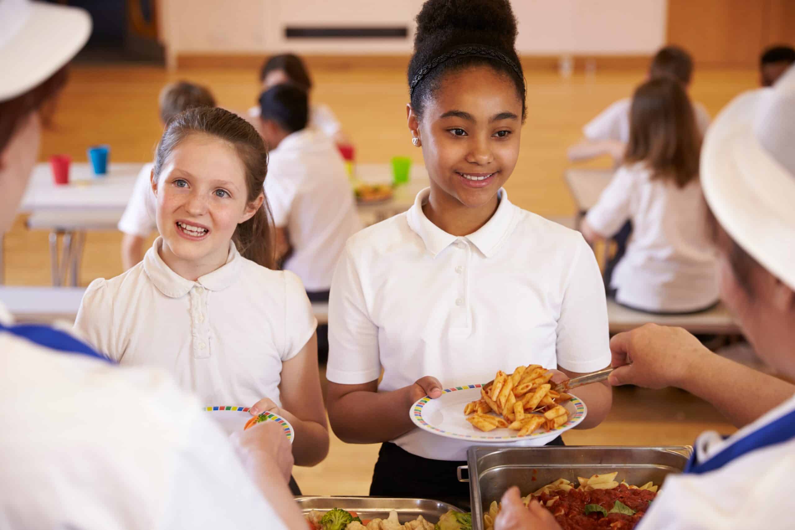 Dos niñas con uniforme escolar sostienen unos platos y sonríen mientras los empleados del comedor les sirven la comida. Otros alumnos están sentados comiendo en las mesas que se ven al fondo.