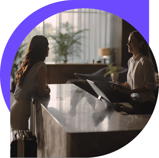 Dos mujeres conversan en la recepción de un hotel; una está de pie con una maleta y la otra está sentada delante de un ordenador, con plantas y una iluminación tenue al fondo. Un semicírculo morado perfila la parte superior de la imagen.