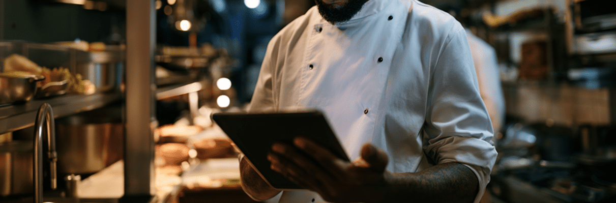 Un chef con uniforme blanco sostiene una tableta mientras se encuentra de pie en la ajetreada cocina de un restaurante, con estanterías y alimentos al fondo.