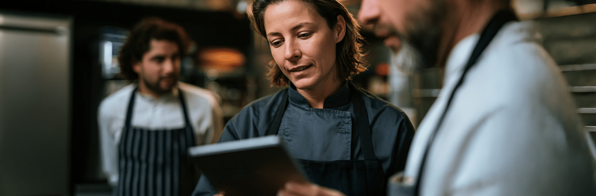 Tres cocineros en una cocina; dos de ellos, en primer plano, están hablando de algo mientras miran una tableta, mientras que el tercero está de pie al fondo, observando.