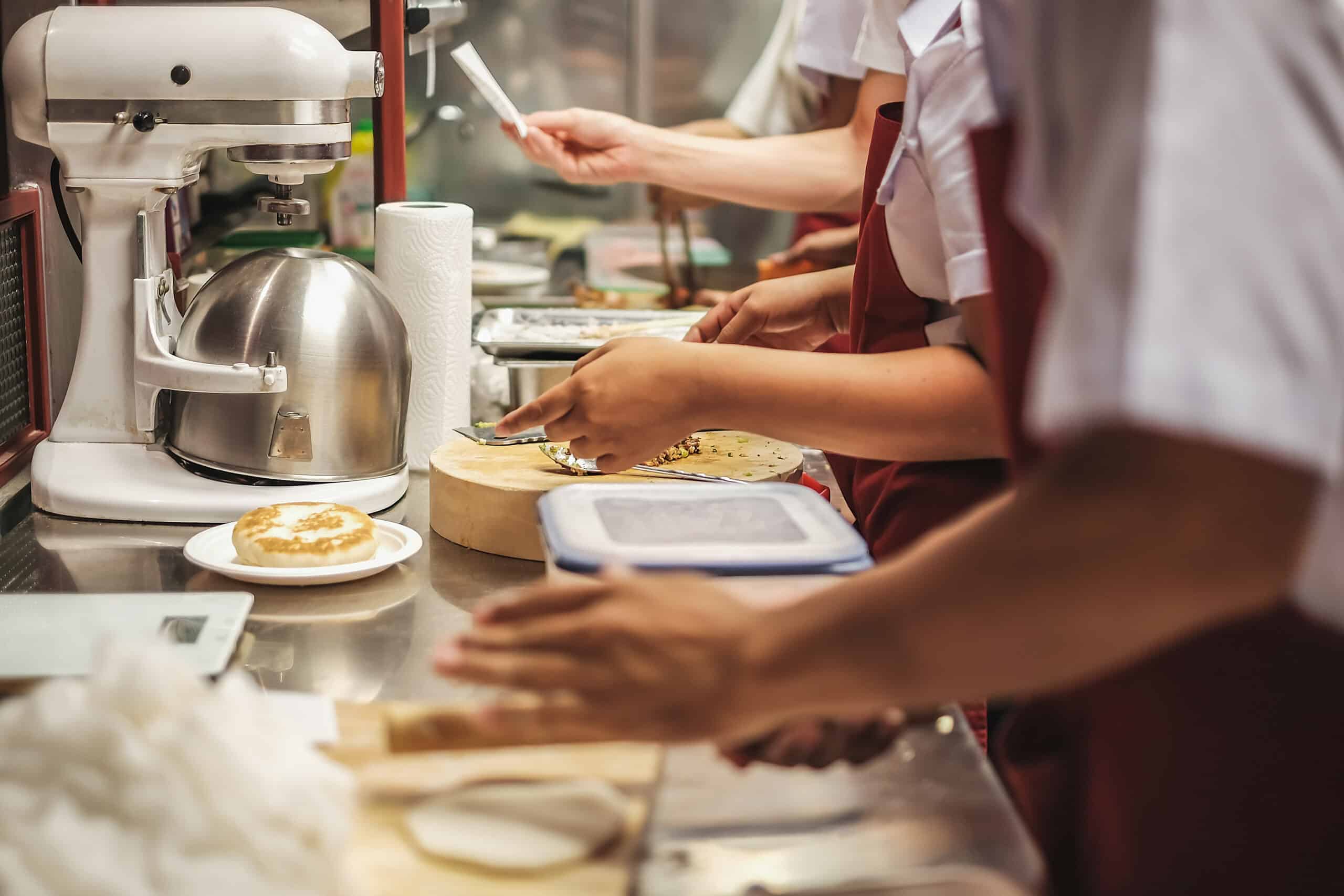 Varias personas con delantal preparan comida en una encimera de cocina de acero inoxidable. En el ajetreado espacio de trabajo se ven una batidora de pie, toallas de papel y platos con masa o pastelería.