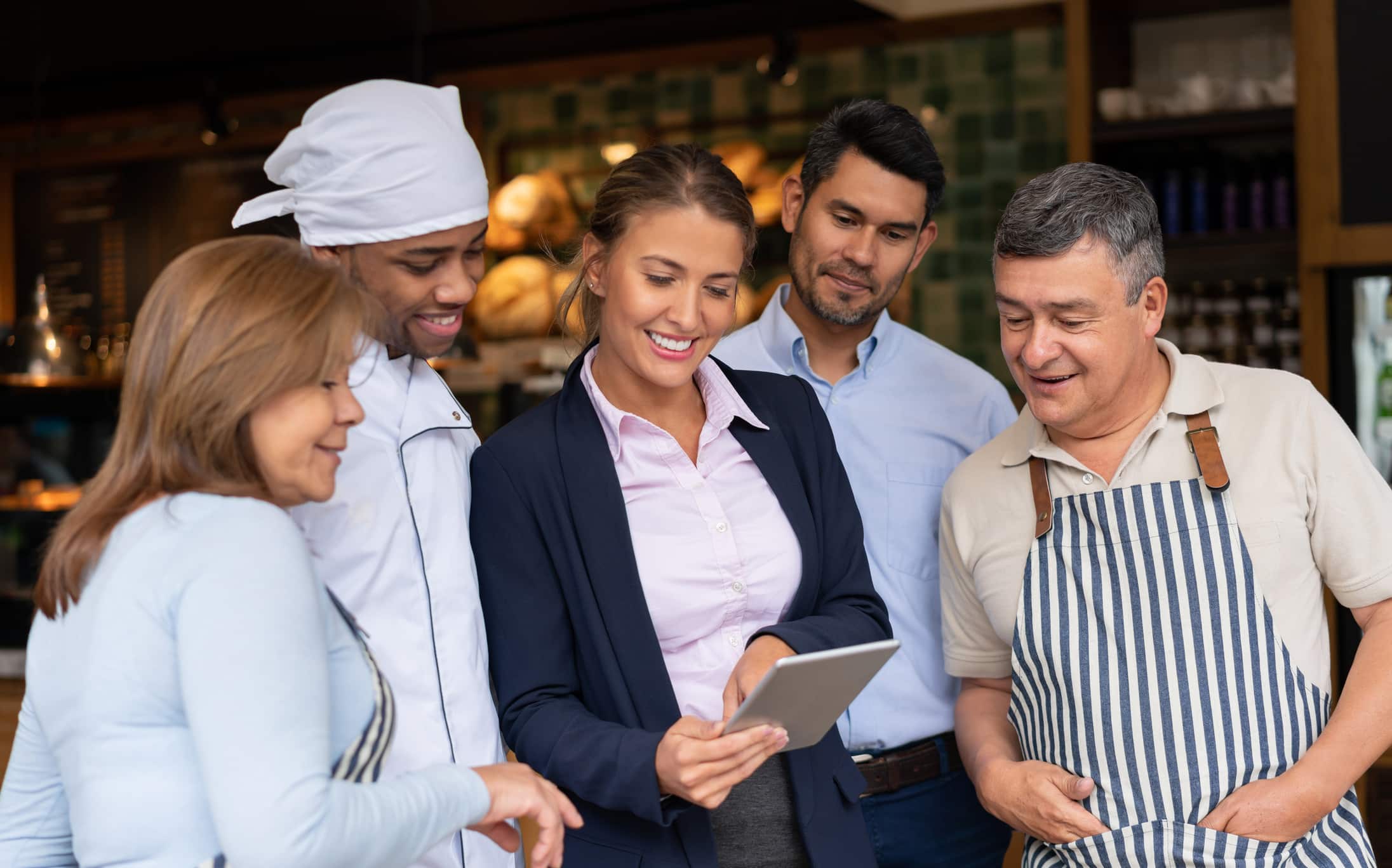 Un grupo de cinco adultos, entre los que hay dos con delantal, sonríen y miran juntos una tableta en un restaurante o una cafetería. Una mujer vestida con traje de negocios sostiene la tableta, mientras los demás se agrupan a su alrededor.