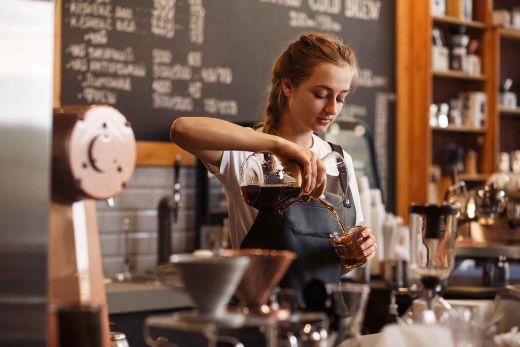 Un barista, que lleva un delantal, sirve café de una jarra de cristal a una taza detrás de la barra de una cafetería, con el equipo de café y una pizarra con el menú al fondo.