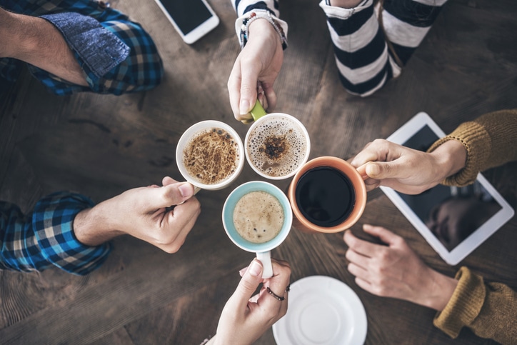 Cuatro personas hacen chocar sus tazas de café sobre una mesa de madera, en la que se ven una tableta, un teléfono y un plato vacío.