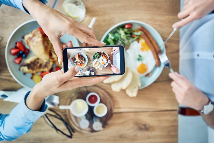 Una persona que está haciendo una foto de los platos del desayuno con un smartphone; en los platos hay huevos, salchichas, ensalada, tostadas y fruta sobre una mesa de madera, mientras otra persona sostiene un tenedor y un cuchillo, lista para comer.