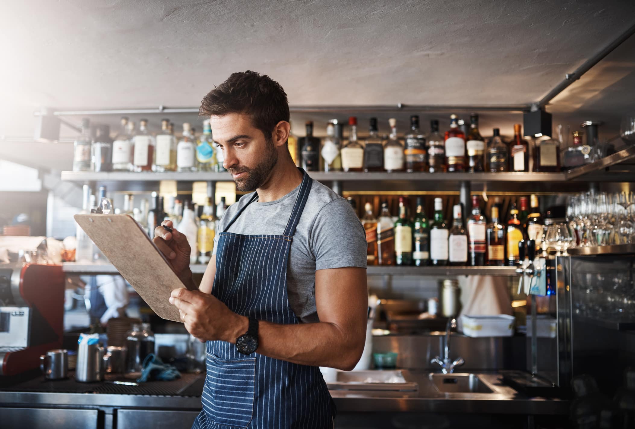 Un hombre que lleva un delantal está de pie en un bar o restaurante, escribiendo en una carpeta con estantes llenos de botellas y vasos a sus espaldas.