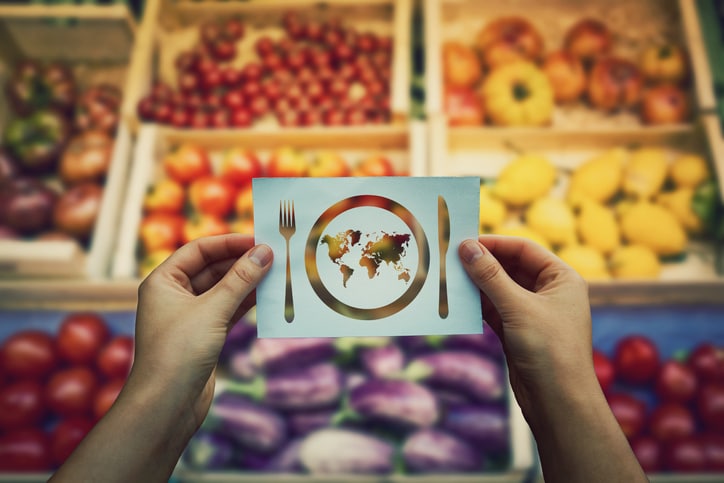 Una persona sostiene una tarjeta en la que se ve un plato con un mapamundi, un tenedor y un cuchillo, con una variedad de frutas y verduras difuminadas al fondo.