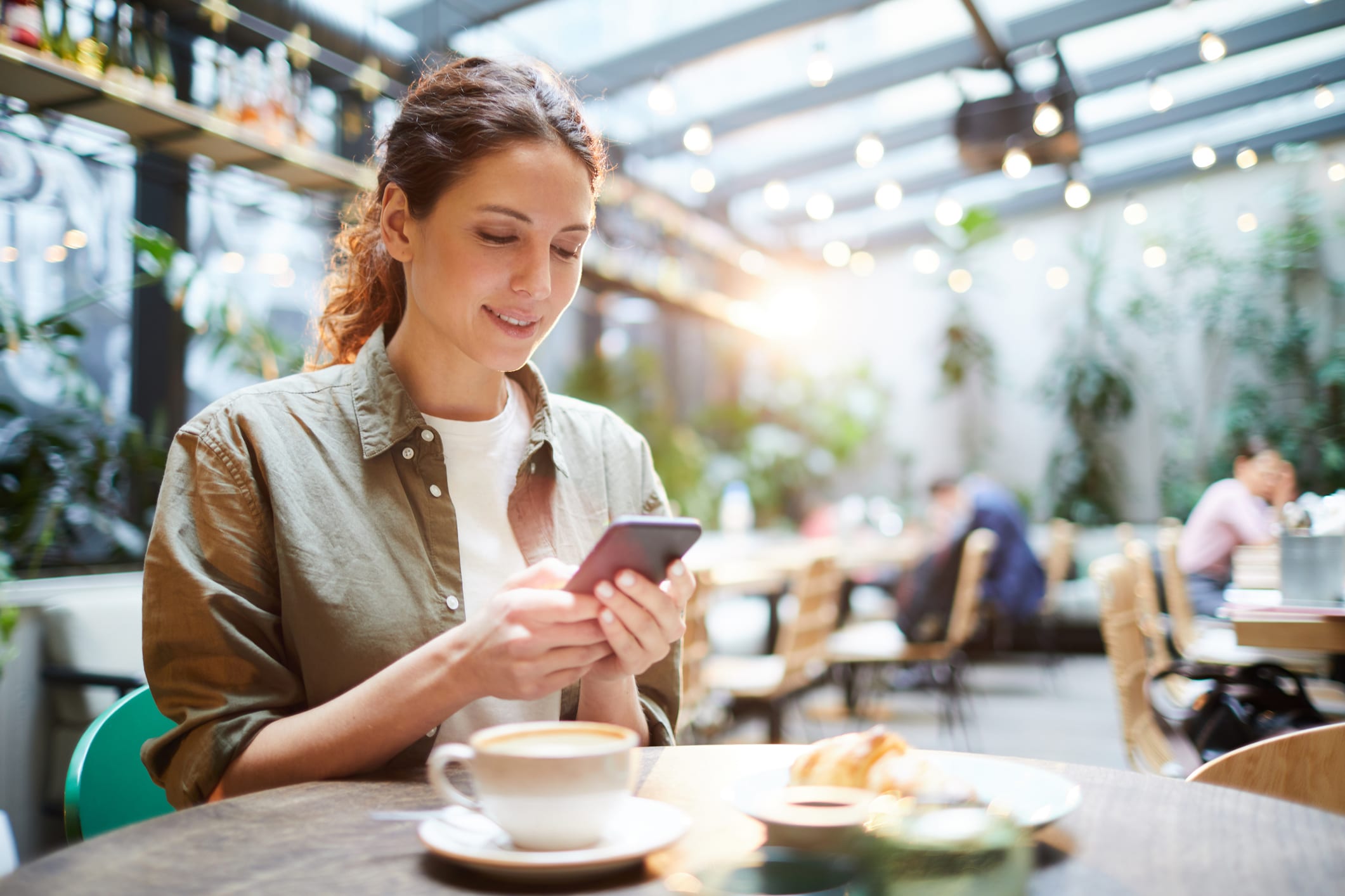 Una mujer está sentada en la mesa de una cafetería, sonriendo mientras usa su móvil. Delante de ella hay una taza de café y un bollo. El fondo es luminoso, con lámparas colgantes y plantas verdes.