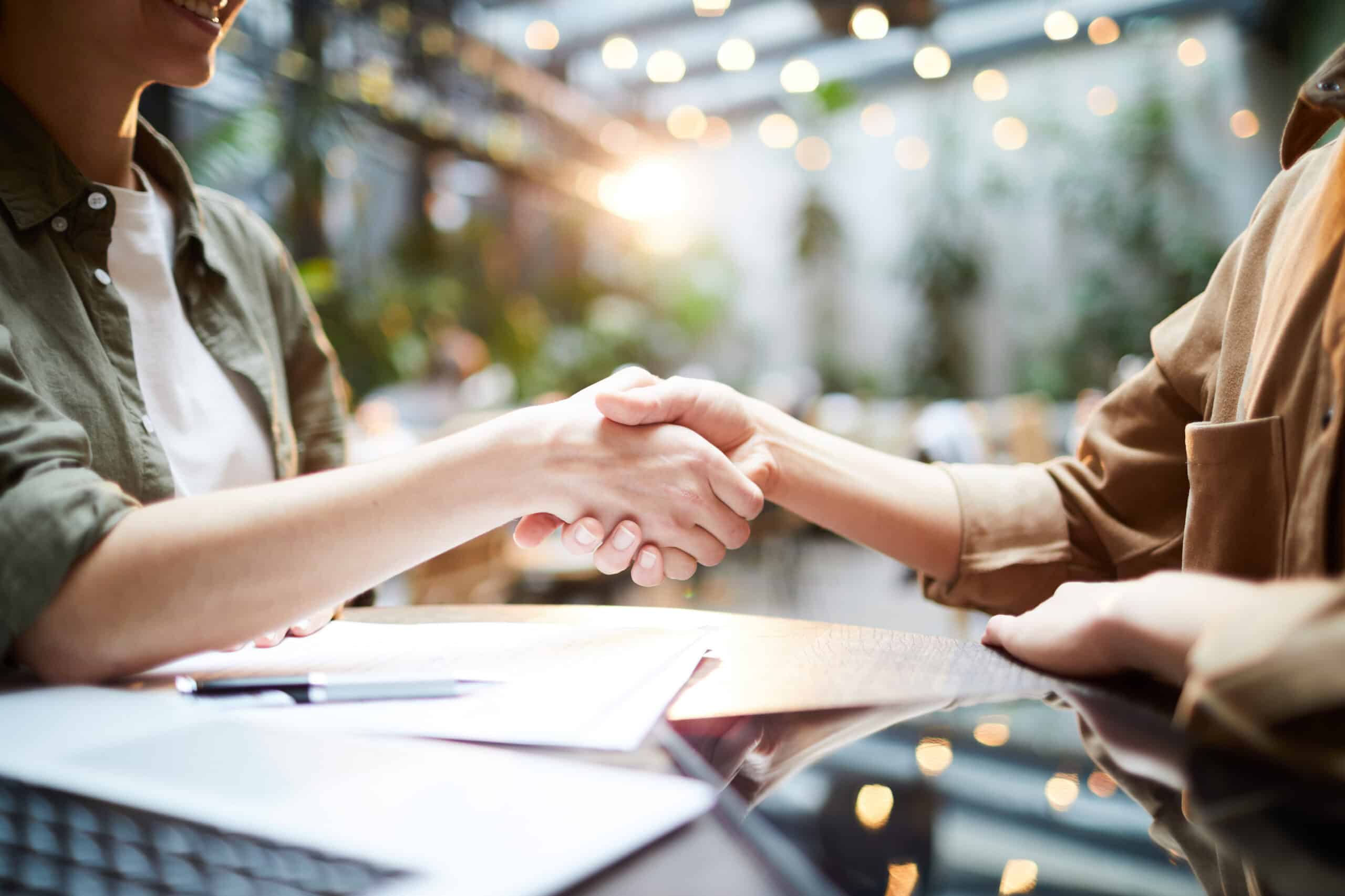 Dos personas se dan la mano al otro lado de una mesa en un entorno interior luminoso y moderno, lo que sugiere una reunión o un acuerdo satisfactorio.