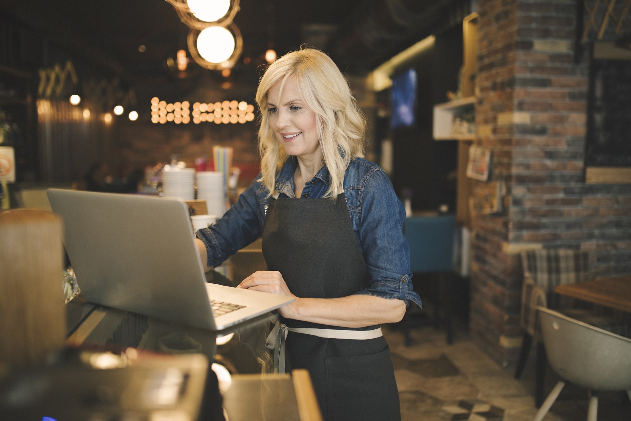 Una mujer que lleva un delantal negro y una camisa vaquera trabaja con un ordenador portátil detrás de la barra de una cafetería, con luces cálidas y un interior acogedor y moderno al fondo.
