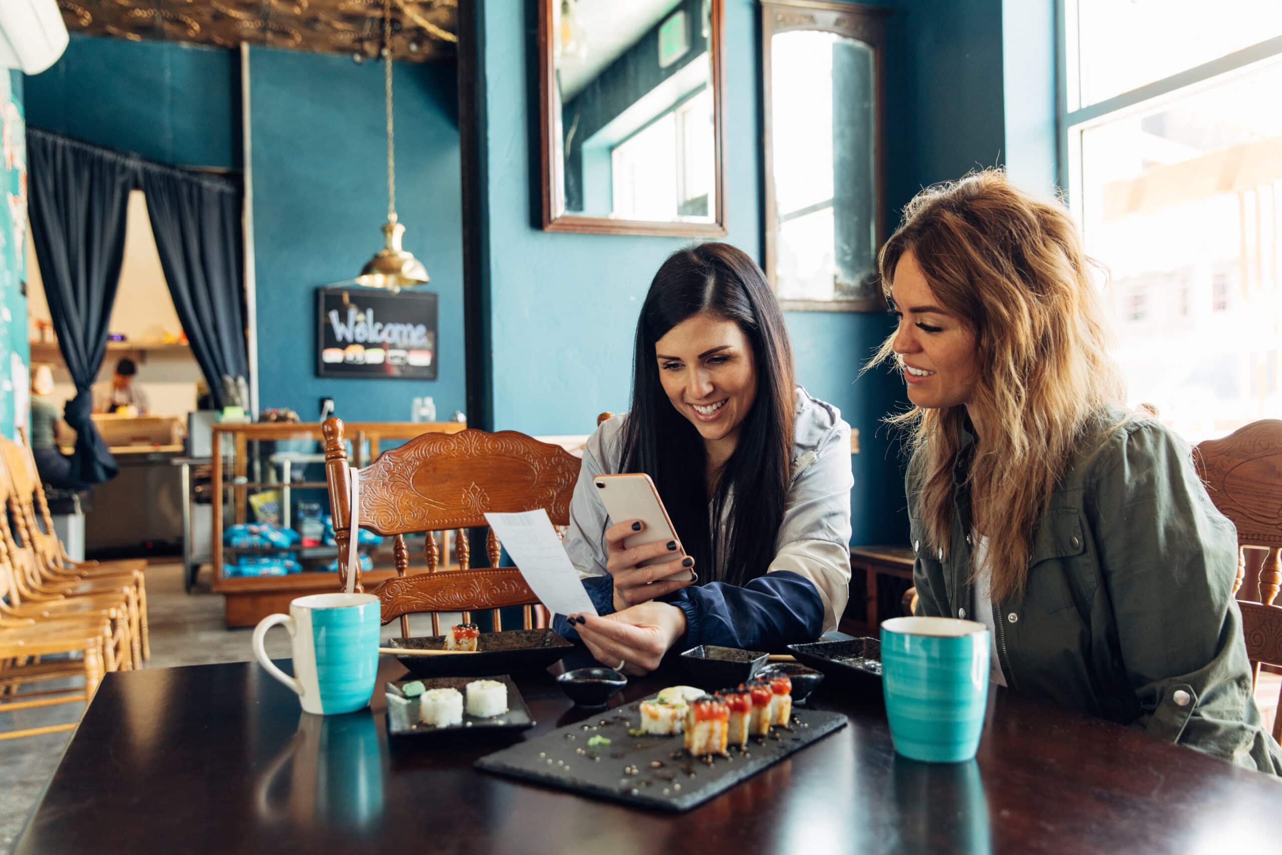 Dos mujeres están sentadas en una mesa de una cafetería con tazas de café y sushi, sonriendo y mirando juntas un móvil y un recibo. La cafetería tiene paredes azules y sillas de madera.