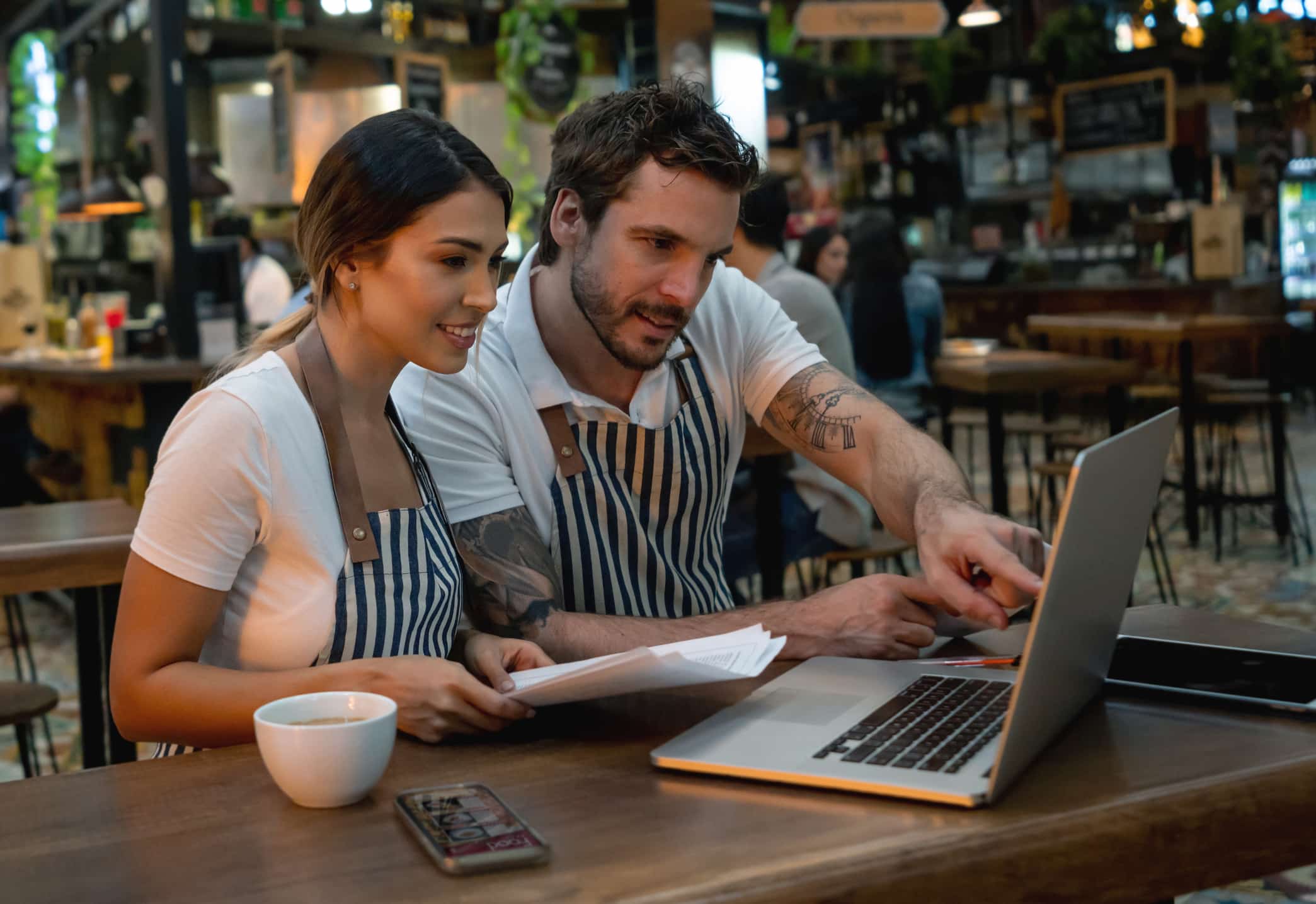Dos personas con delantal están sentadas en la mesa de una cafetería, mirando un ordenador portátil y revisando unos documentos. Sobre la mesa hay una taza de café y un smartphone. Al fondo se ve el ambiente informal y animado de una cafetería o un restaurante.