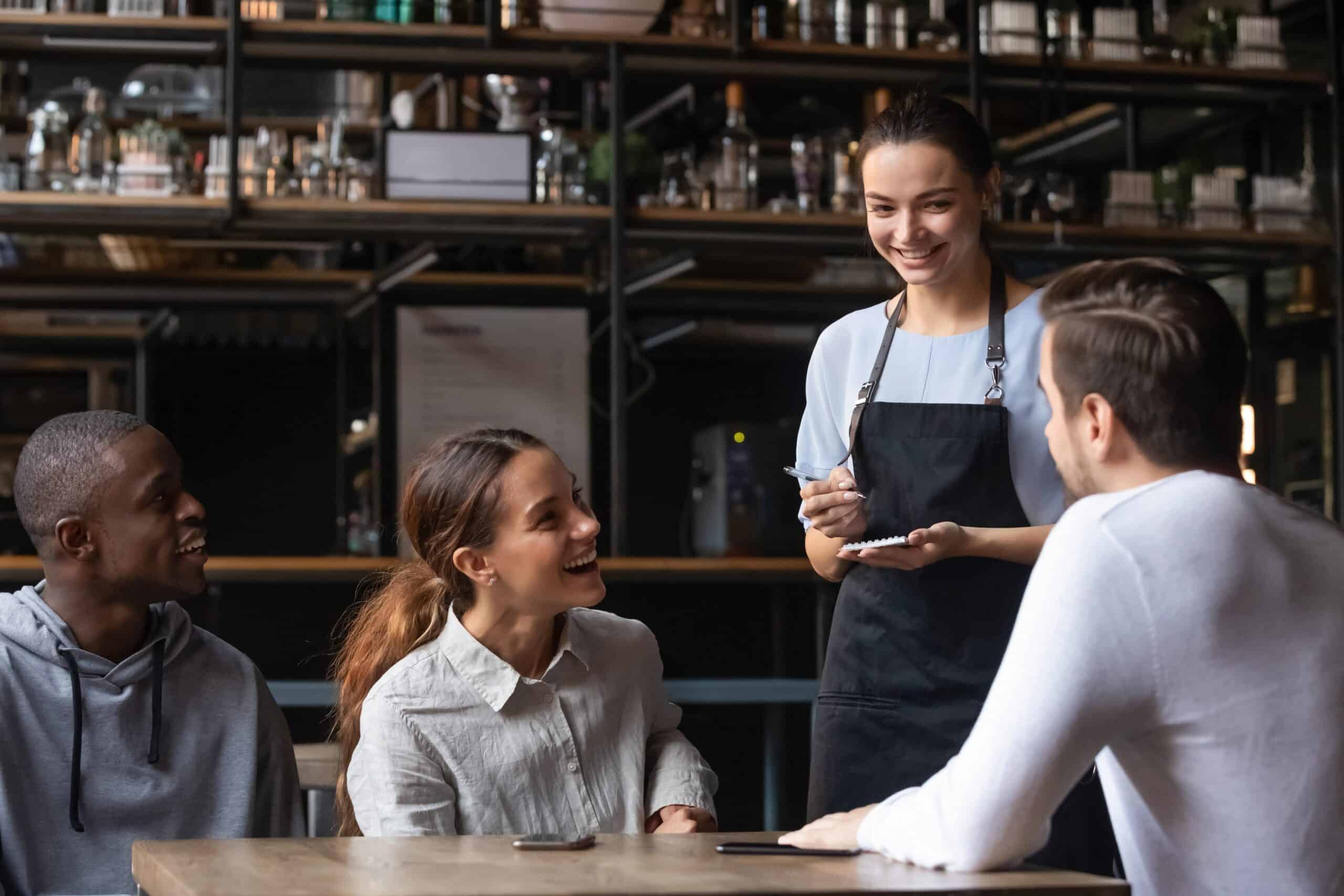 Tres personas están sentadas en una mesa, sonriendo y hablando con una camarera que se encuentra de pie junto a ellas, con un bloc de notas y un bolígrafo en la mano, lista para tomarles la comanda en una acogedora cafetería o restaurante.