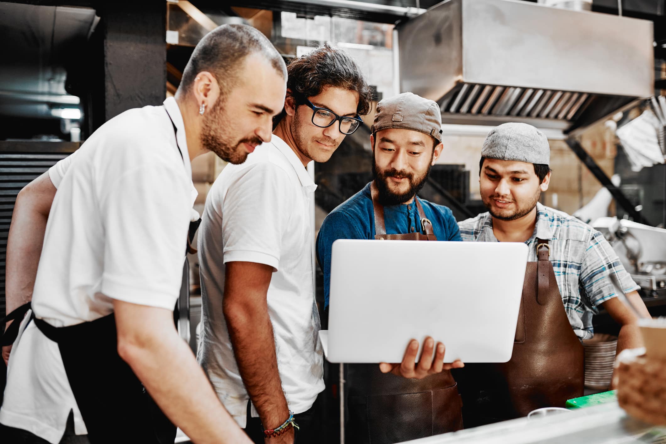 Cuatro personas con delantales están de pie en una cocina, mirando juntas la pantalla de un ordenador portátil y sonriendo, rodeadas de electrodomésticos de acero inoxidable y utensilios de cocina.