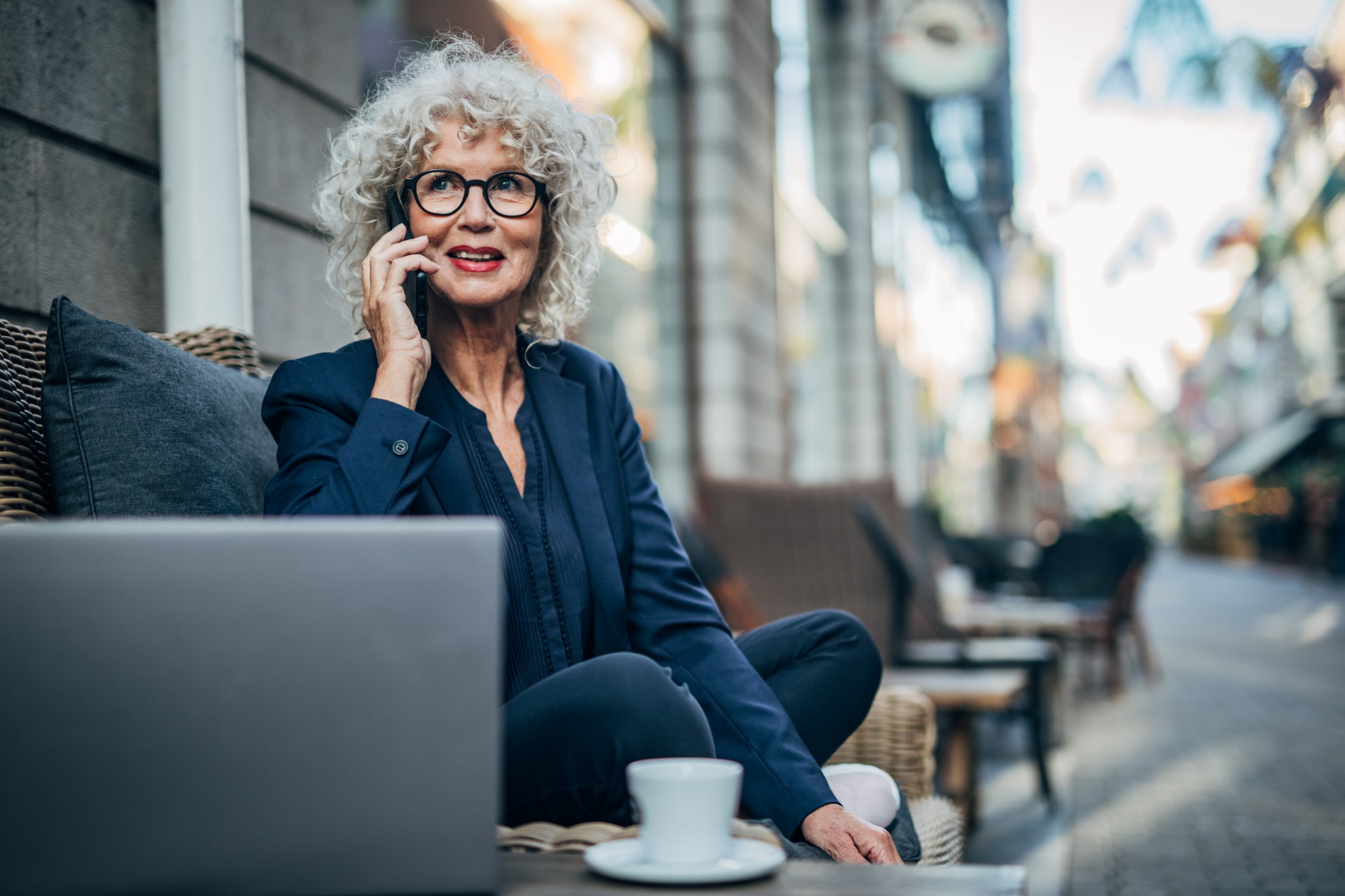 Una mujer mayor, sonriente, con el pelo gris y rizado y gafas, está sentada al aire libre en la mesa de una cafetería, hablando por el móvil. Delante de ella hay un ordenador portátil y una taza de café. Al fondo se ve una calle de la ciudad.