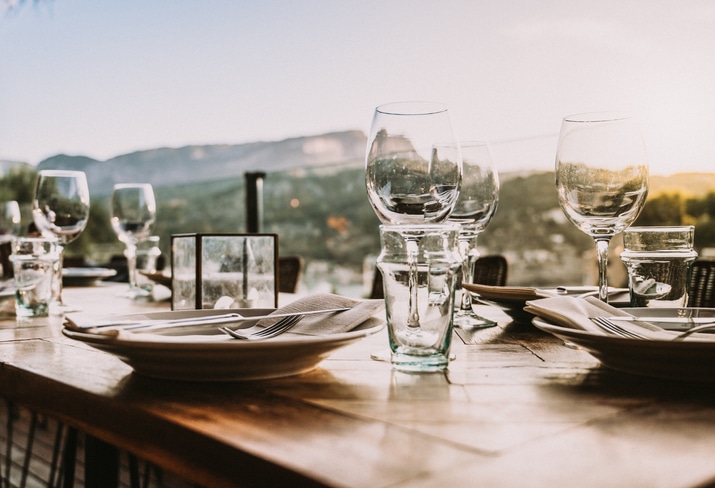 Primer plano de una mesa puesta con platos, tenedores, vasos y servilletas al aire libre durante la puesta de sol, con vistas a unas montañas difuminadas y un paisaje pintoresco al fondo.