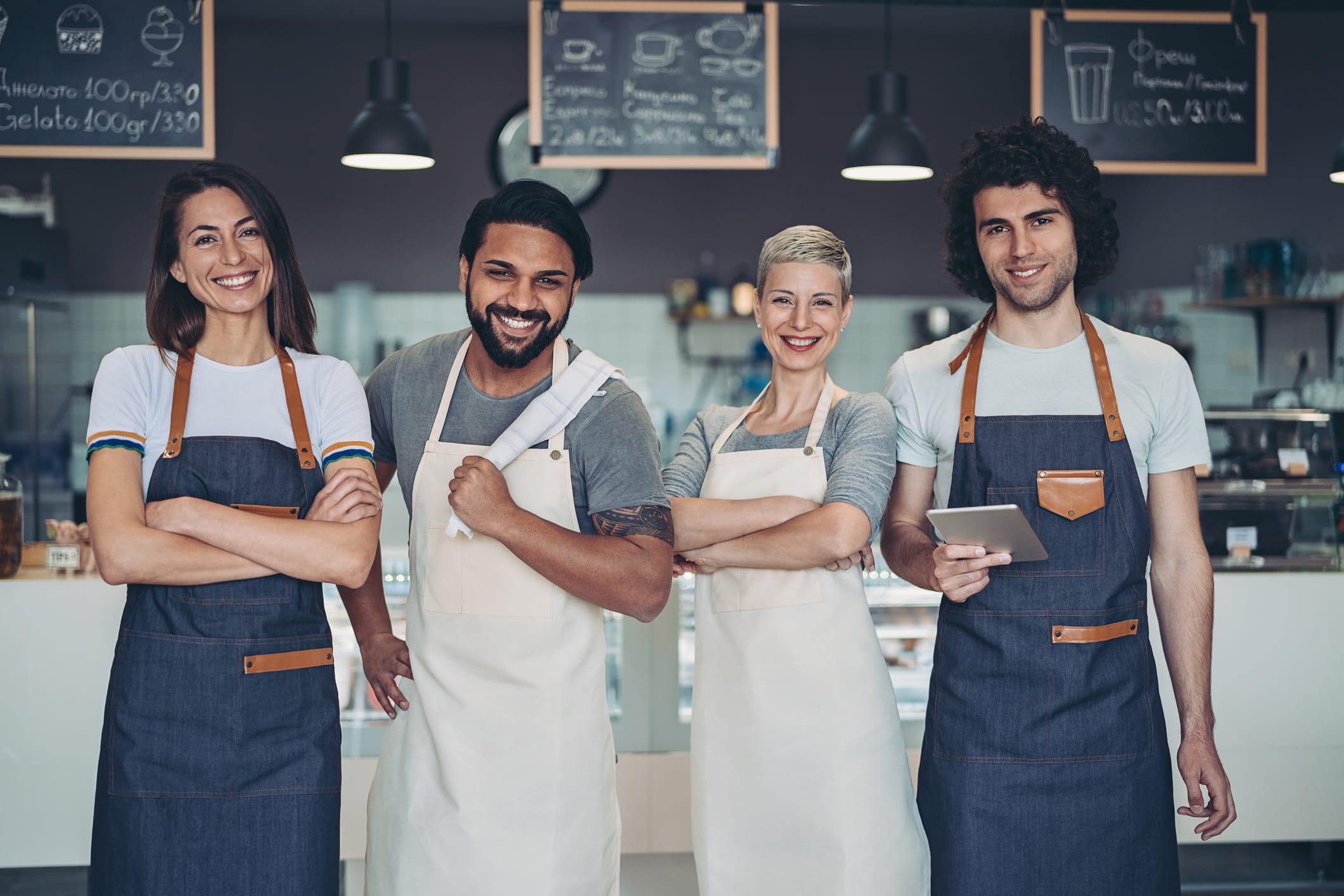 Cuatro empleados de una cafetería, sonrientes y con delantales, están de pie juntos delante de una barra, con los brazos cruzados o sosteniendo una tableta, y con menús escritos en pizarras colgados al fondo.
