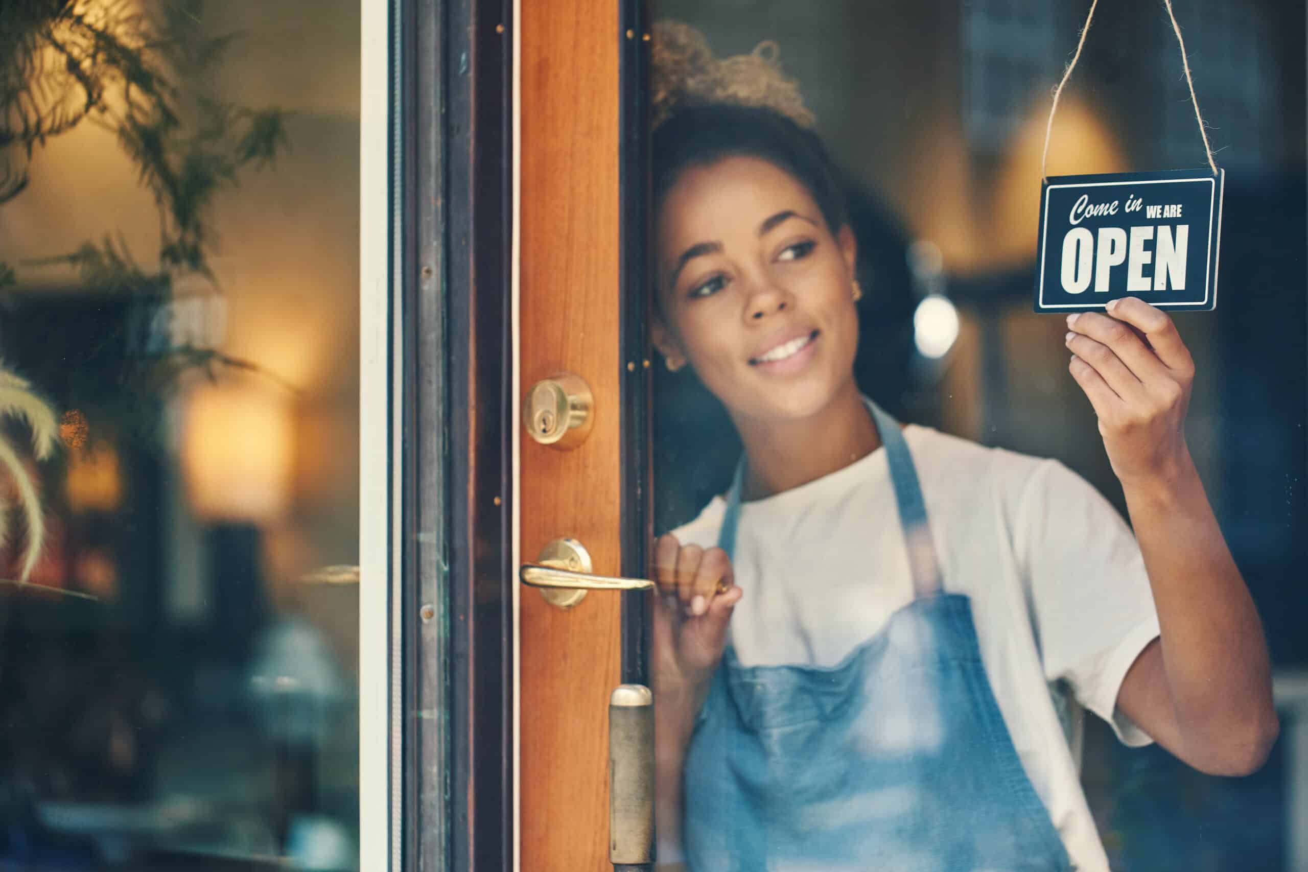 Una persona sonriente, con un delantal puesto, cuelga un cartel de «Abierto» en la puerta de cristal de una tienda, preparándose para dar la bienvenida a los clientes.
