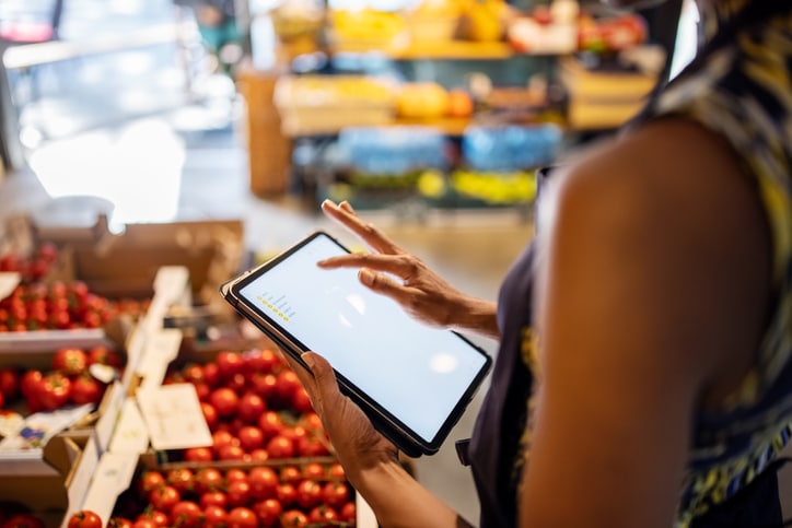 Una persona utiliza una tableta junto a unas cajas de tomates en un mercado o una tienda de alimentación, quizá para comprobar el inventario o gestionar datos.