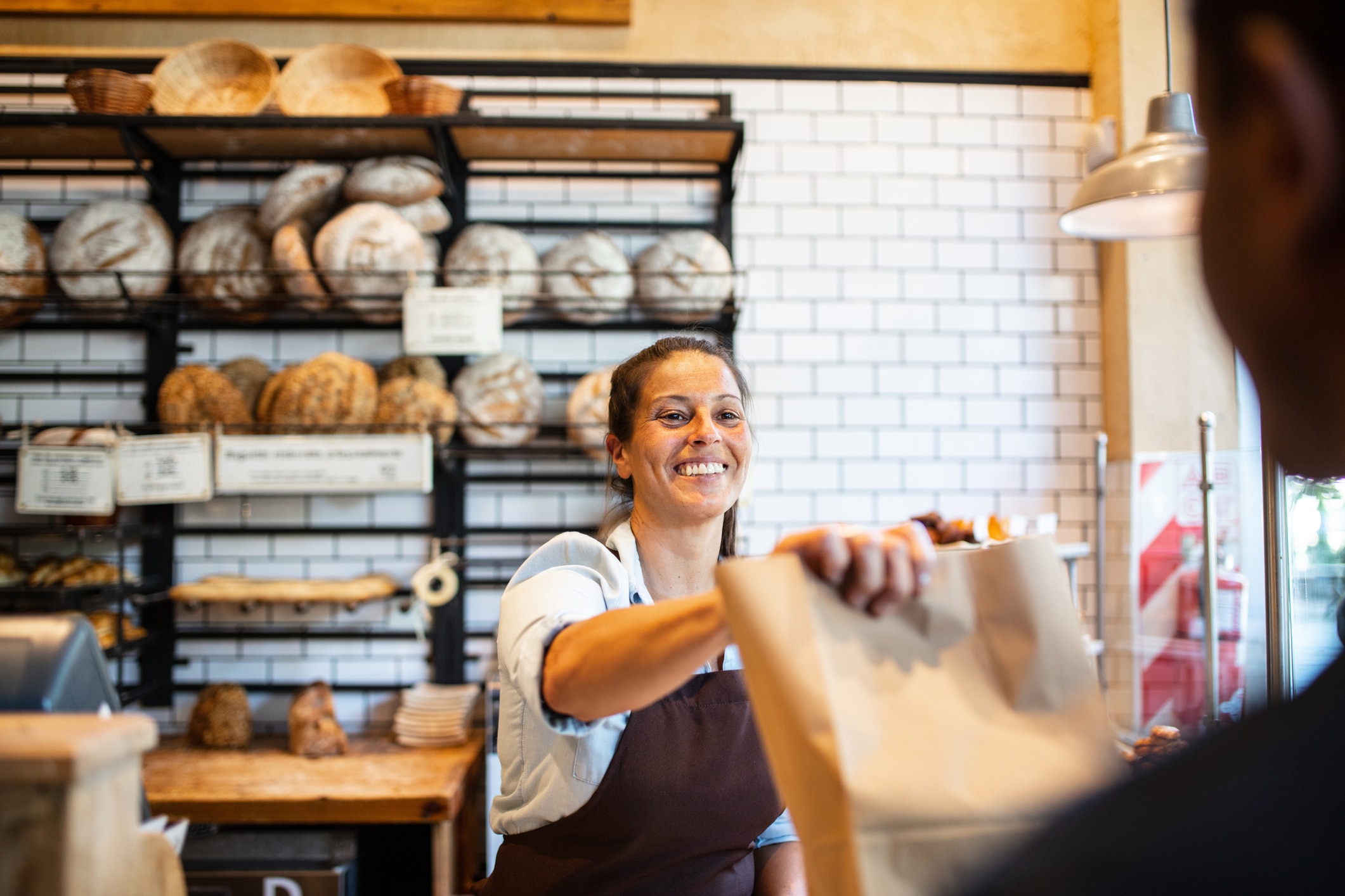 Un empleado de panadería, sonriente y con delantal, le entrega una bolsa de papel a un cliente. Al fondo se ven estanterías con diferentes tipos de pan y una pared de azulejos blancos.