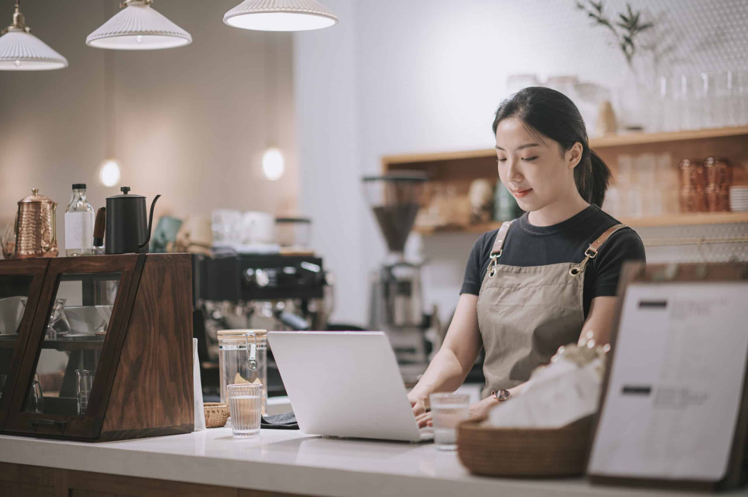 Un barista, que lleva un delantal, trabaja con un ordenador portátil detrás del mostrador de una cafetería, rodeado de utensilios de café, cristalería y una iluminación cálida.