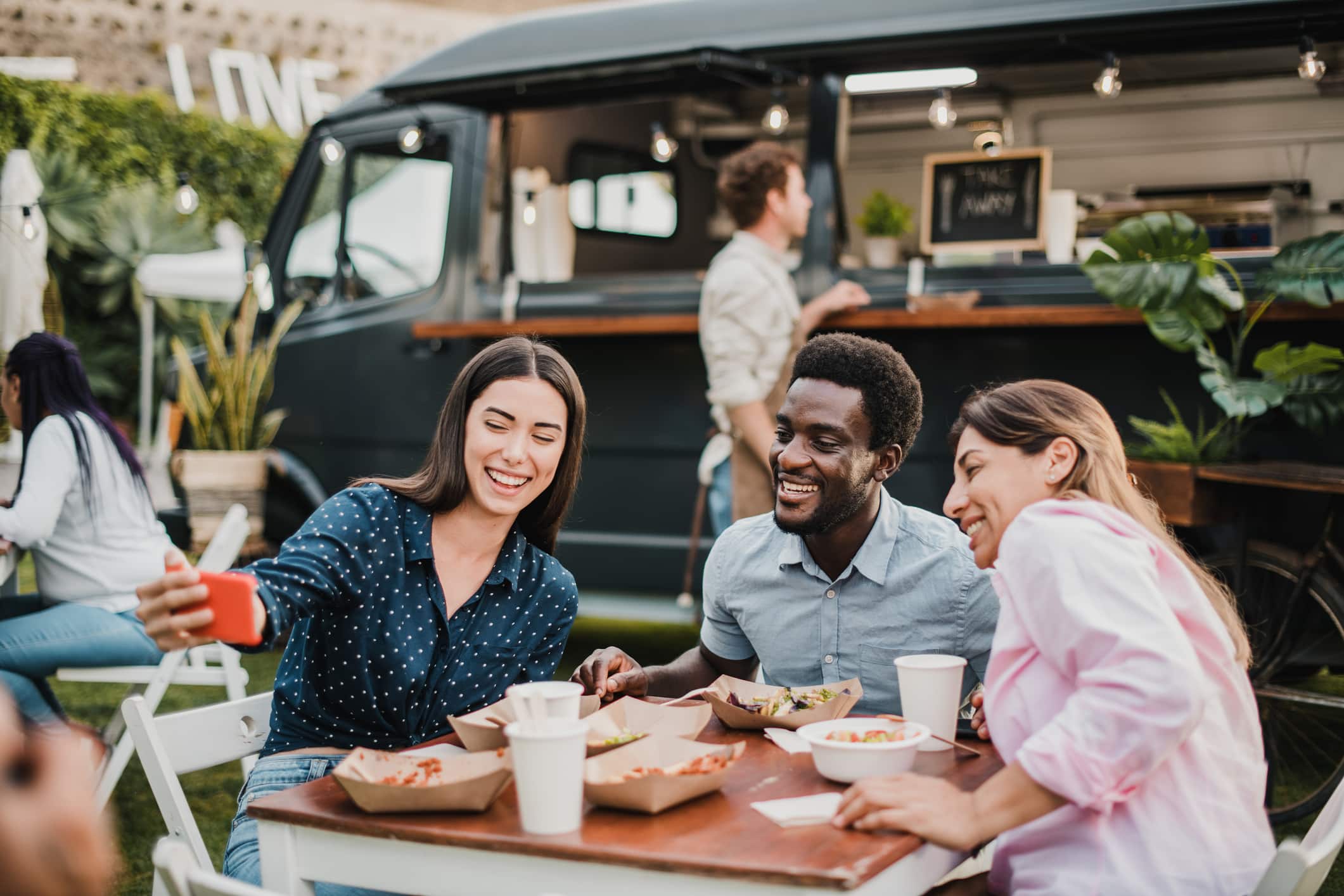 Tres personas están sentadas en una mesa al aire libre con comida, sonriendo y haciéndose un selfi juntas delante de un food truck negro en un día soleado.