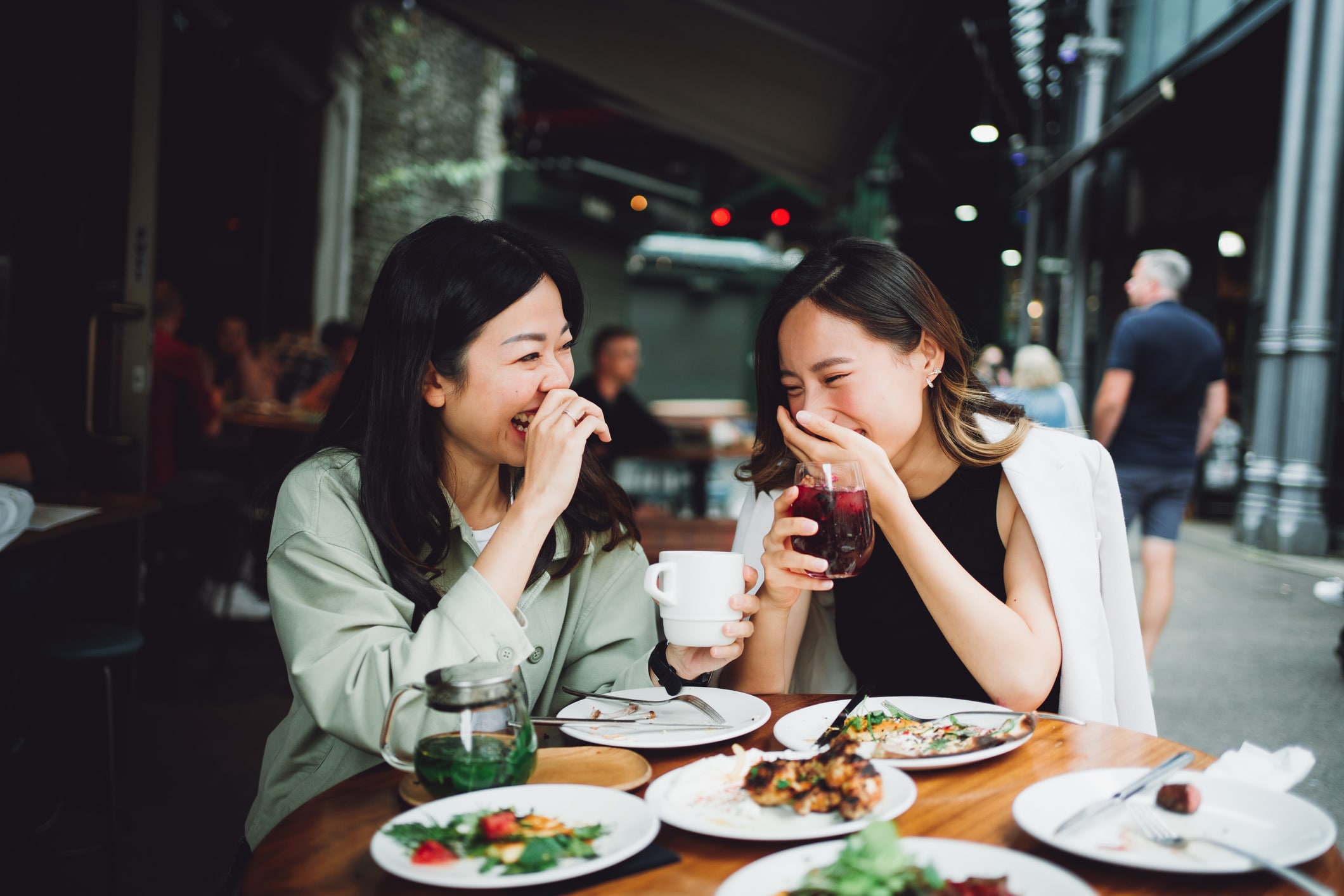 Dos mujeres están sentadas en la terraza de una cafetería, riendo y compartiendo bebidas y comida. Sobre la mesa hay platos de ensalada, pan y bebidas. Al fondo se ven otros comensales y una calle de la ciudad.
